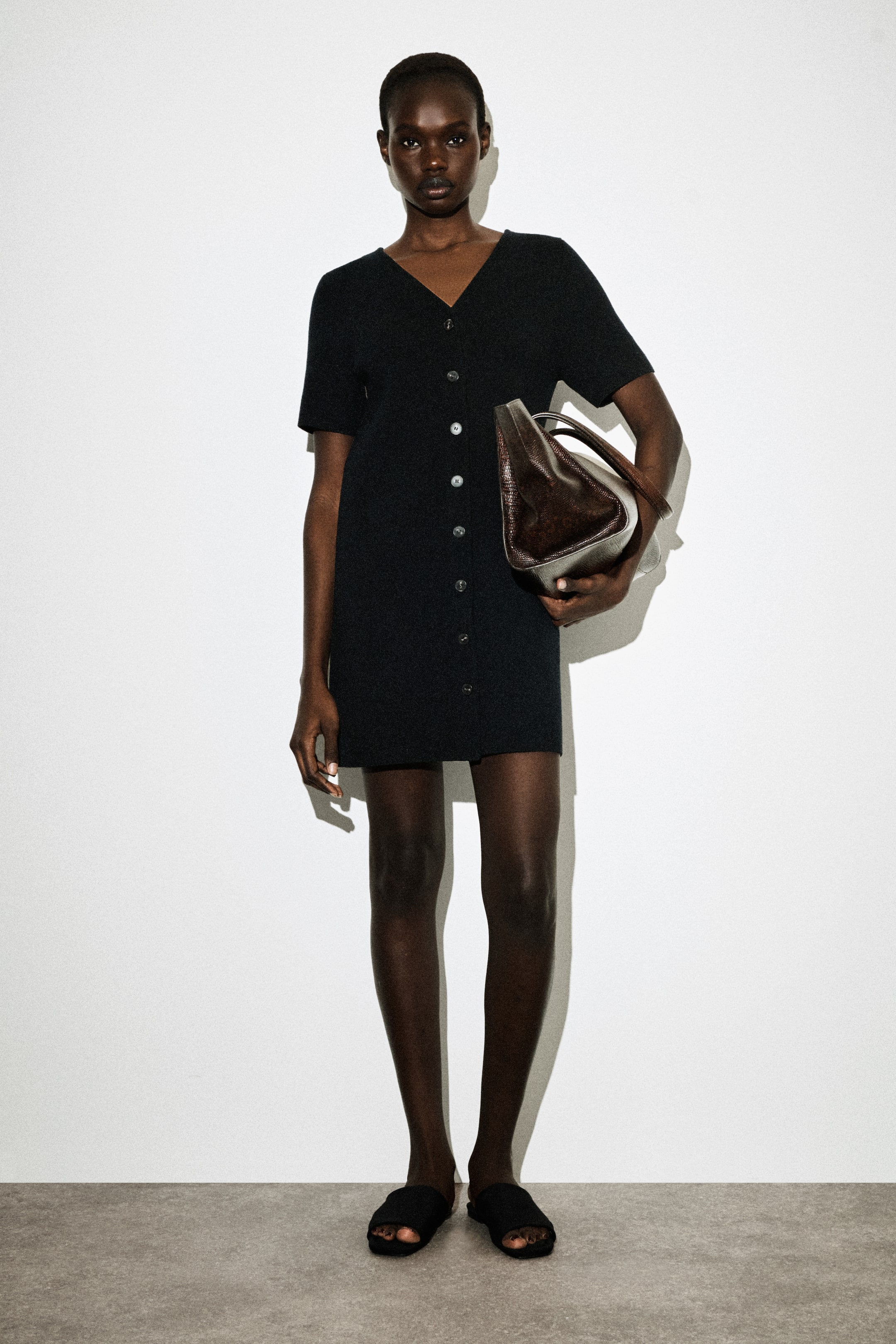 Größeres Bild ansehen: Ein schwarzes Mini-Kleid mit V-Ausschnitt wird von vorne gezeigt, mit kurzen Ärmeln und einer vollständigen Knopfleiste entlang des strukturierten Strickstoffs.