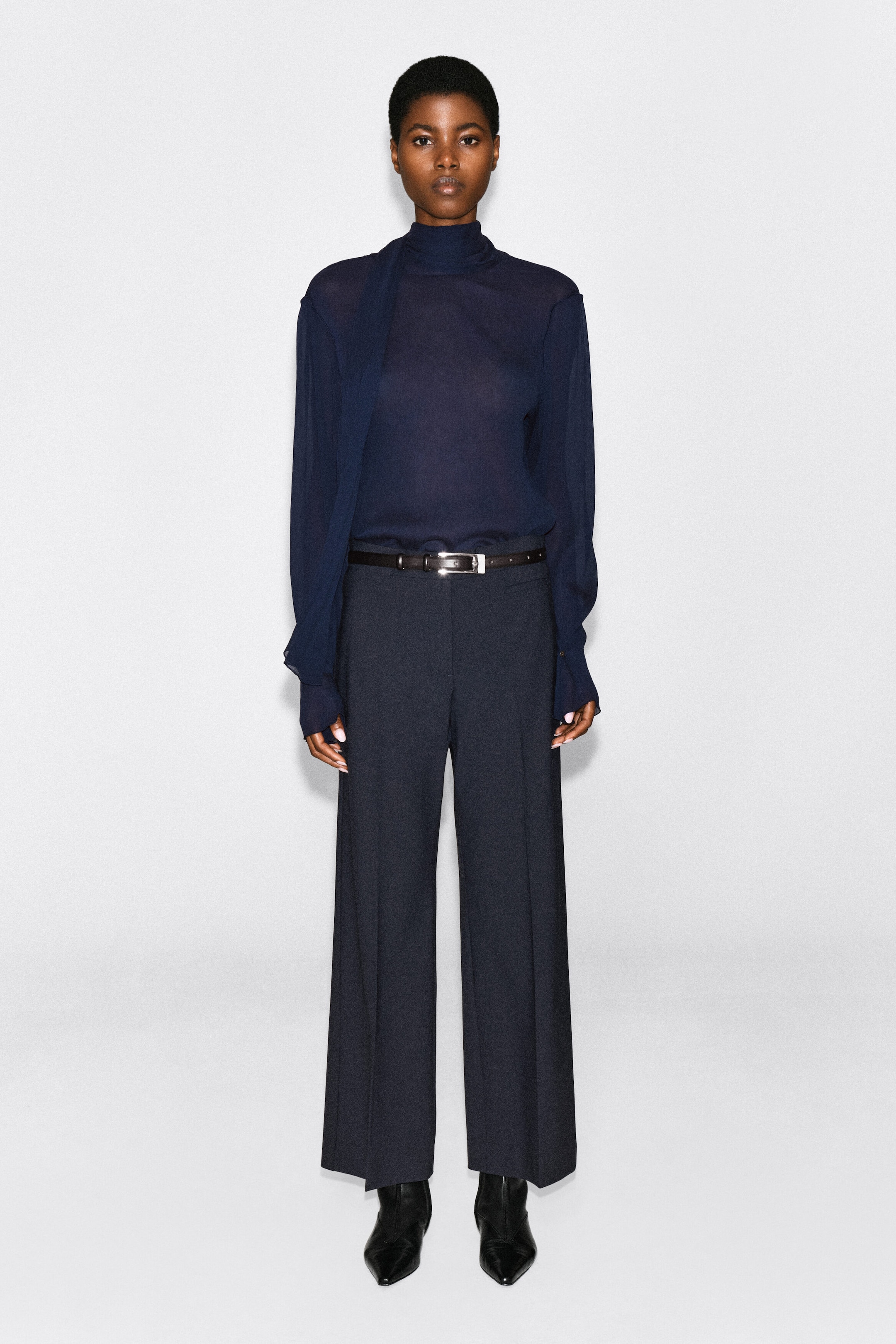 A semi-sheer, dark navy top featuring a high neck and a matching draped scarf is being worn by the model. The long-sleeved top is tucked into dark, wide-leg trousers secured with a black belt. A semi-sheer, dark navy top featuring a high neck and a matching draped scarf is being worn by the model. The long-sleeved top is tucked into dark, wide-leg trousers secured with a black belt.