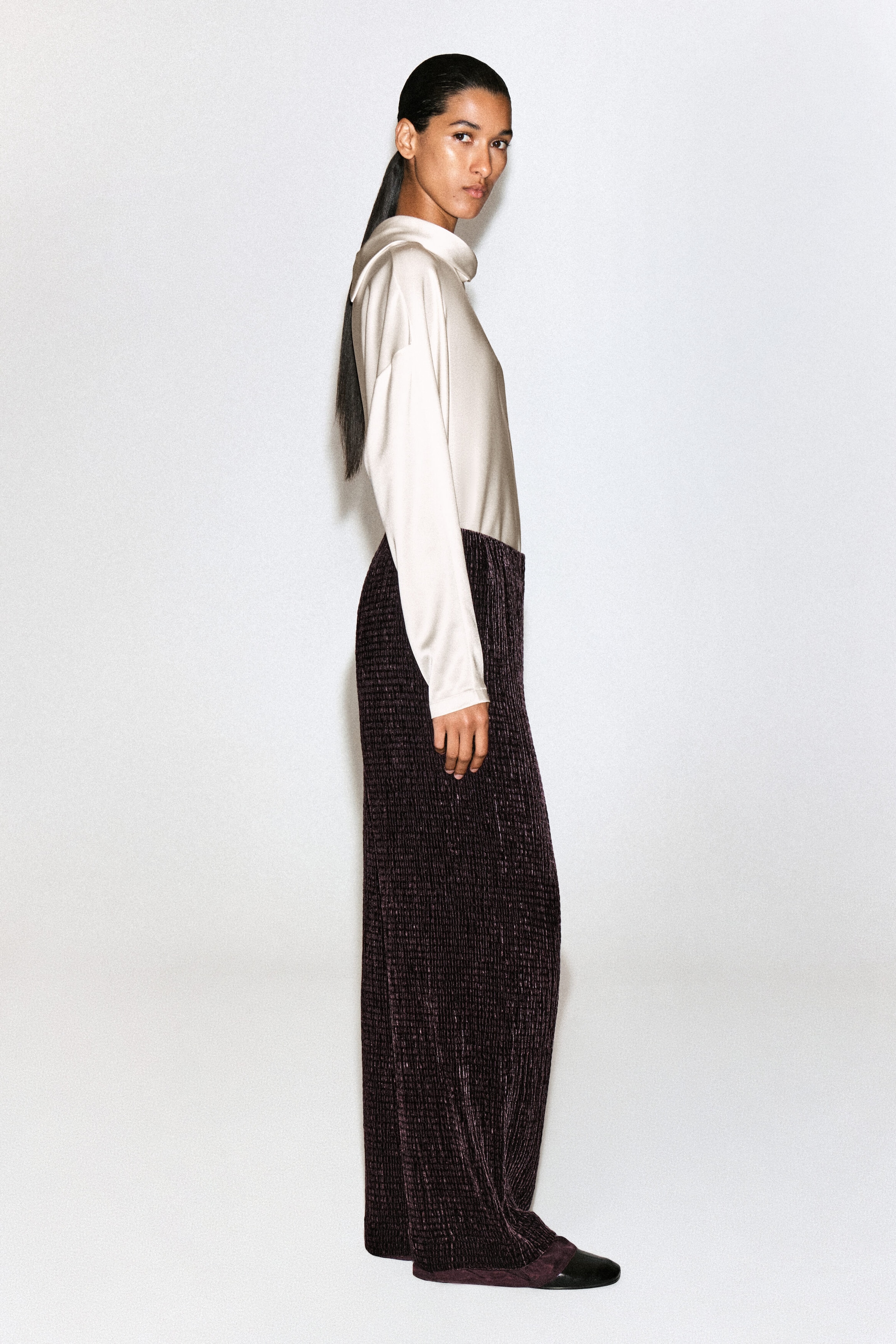 A cream-colored satin blouse with a draped cowl neck and long sleeves is styled tucked into dark purple, textured wide-leg trousers. The ensemble is shown in right-facing side profile, highlighting the fluid drape of the satin top and the crinkled vertical pleats of the trousers. A cream-colored satin blouse with a draped cowl neck and long sleeves is styled tucked into dark purple, textured wide-leg trousers. The ensemble is shown in right-facing side profile, highlighting the fluid drape of the satin top and the crinkled vertical pleats of the trousers.
