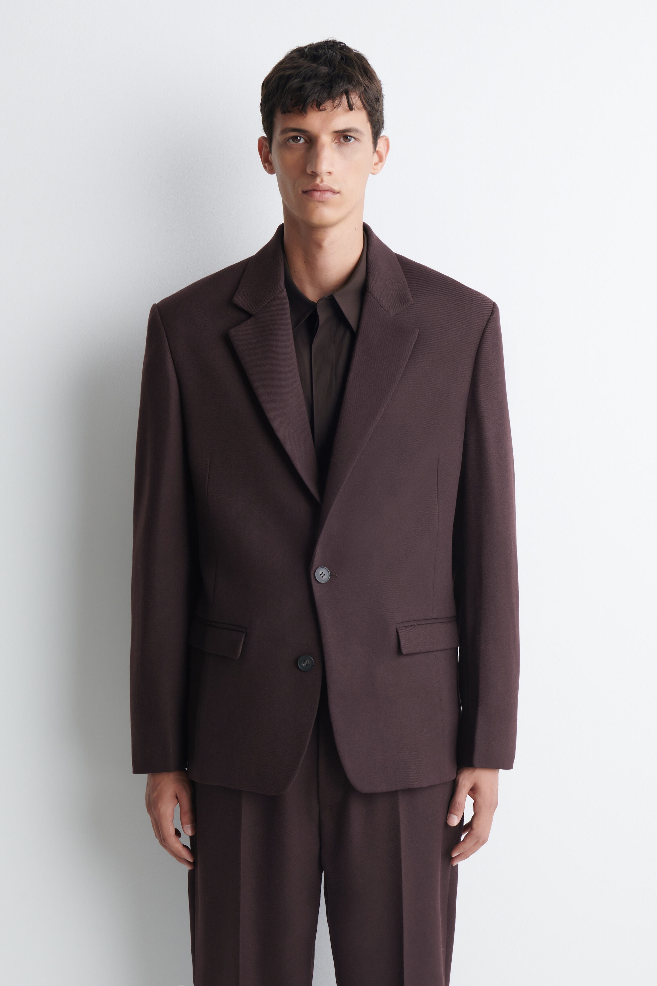 A dark brown suit, comprising a single-breasted blazer with notch lapels and flap pockets, and matching wide-leg trousers, is worn by the model over a dark brown collared shirt. A dark brown suit, comprising a single-breasted blazer with notch lapels and flap pockets, and matching wide-leg trousers, is worn by the model over a dark brown collared shirt.