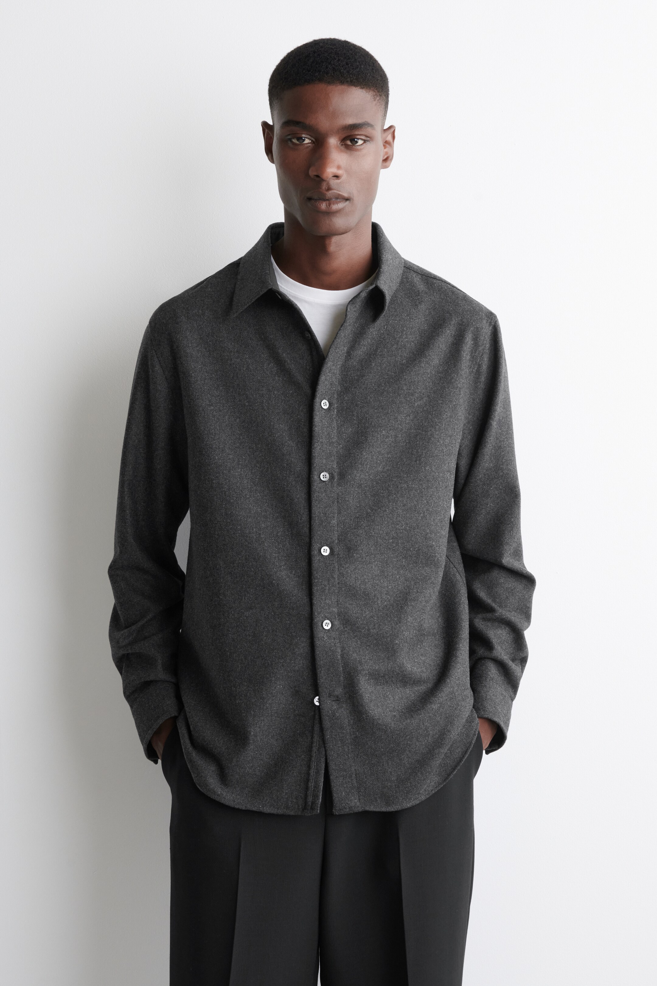 Visualizza immagine più grande: Una camicia grigio scuro con bottoni è presentata da una prospettiva frontale, con un colletto a punta, maniche lunghe e bottoni bianchi lungo la lista frontale. La camicia è indossata sbottonata sopra una t-shirt bianca, mostrando la sua vestibilità rilassata e l'orlo arrotondato.