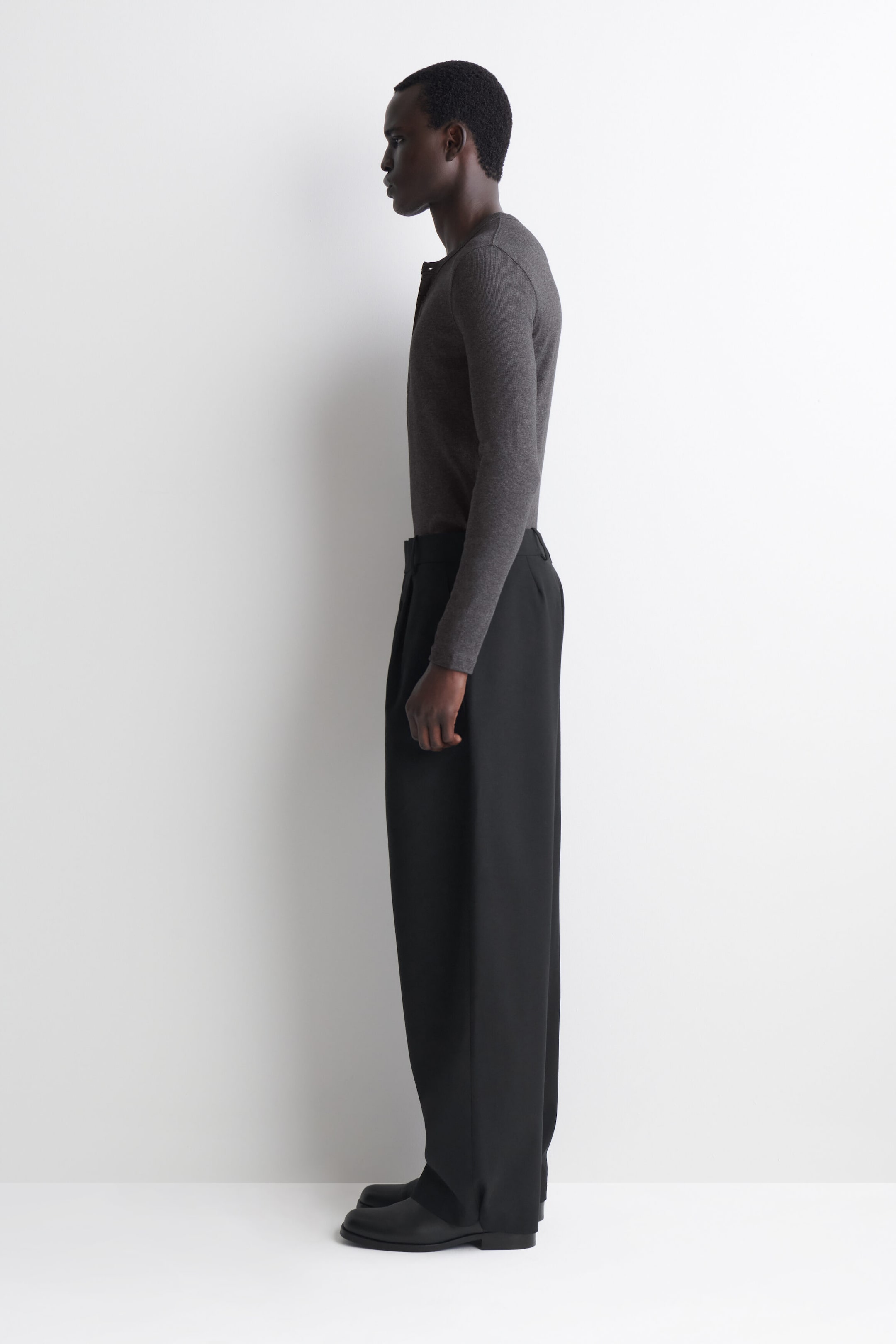 A dark gray slim-fit, ribbed cotton, long-sleeve henley T-shirt is shown in left side profile, highlighting its close fit through the torso and sleeves. The T-shirt, featuring a fine ribbed texture, is tucked into black wide-leg trousers to showcase its streamlined silhouette. A dark gray slim-fit, ribbed cotton, long-sleeve henley T-shirt is shown in left side profile, highlighting its close fit through the torso and sleeves. The T-shirt, featuring a fine ribbed texture, is tucked into black wide-leg trousers to showcase its streamlined silhouette.