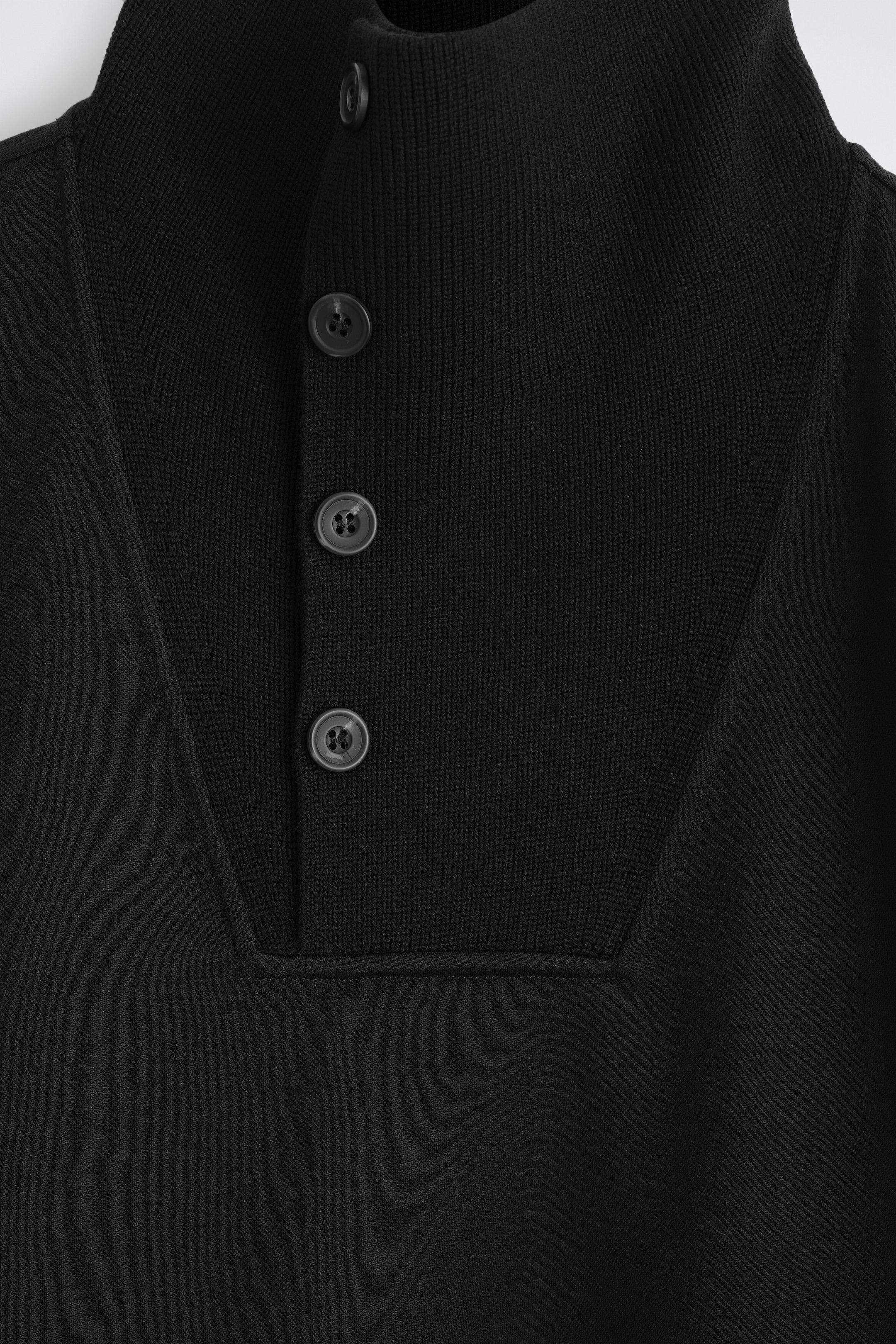 A black garment features a high-neck knit collar and a partial front button placket with four dark gray buttons. The textured knit bib panel contrasts with the smoother fabric of the main body.