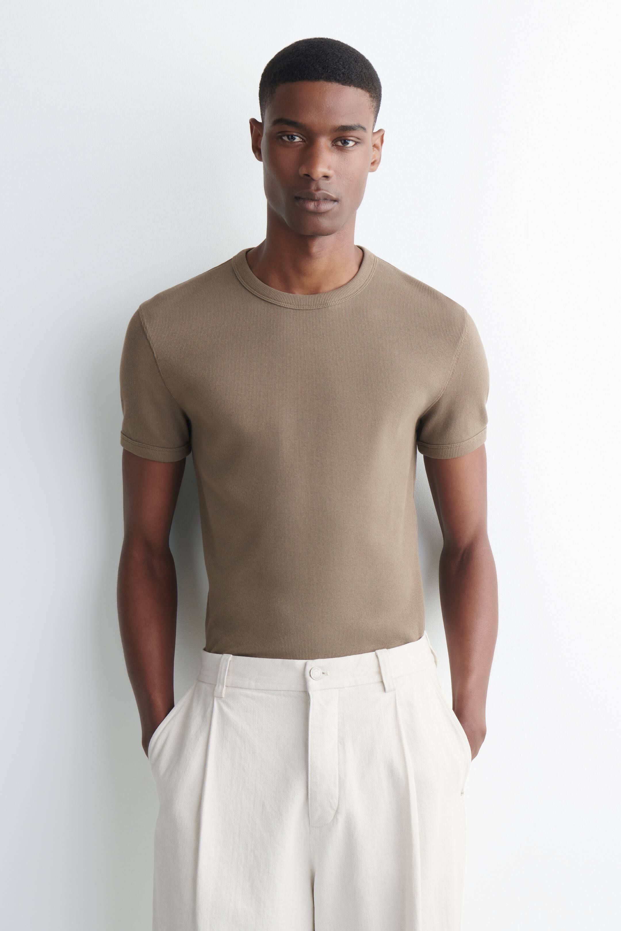 Grösseres Bild ansehen: Ein braunes geripptes T-Shirt mit schmaler Passform, kurzen Ärmeln und Rundhalsausschnitt wird vom Model getragen. Das T-Shirt ist in hellfarbene, plissierte Hosen gesteckt.