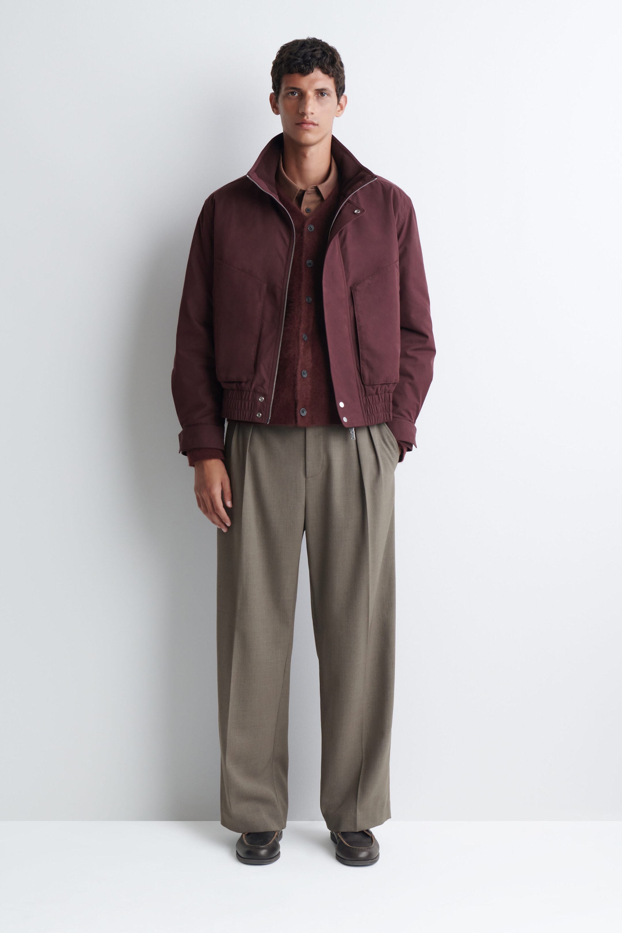 A brushed cashmere V-neck cardigan in deep burgundy is being worn by the model, buttoned over a light brown collared shirt. The cardigan is layered beneath an open burgundy jacket and paired with wide-leg taupe trousers. A brushed cashmere V-neck cardigan in deep burgundy is being worn by the model, buttoned over a light brown collared shirt. The cardigan is layered beneath an open burgundy jacket and paired with wide-leg taupe trousers.