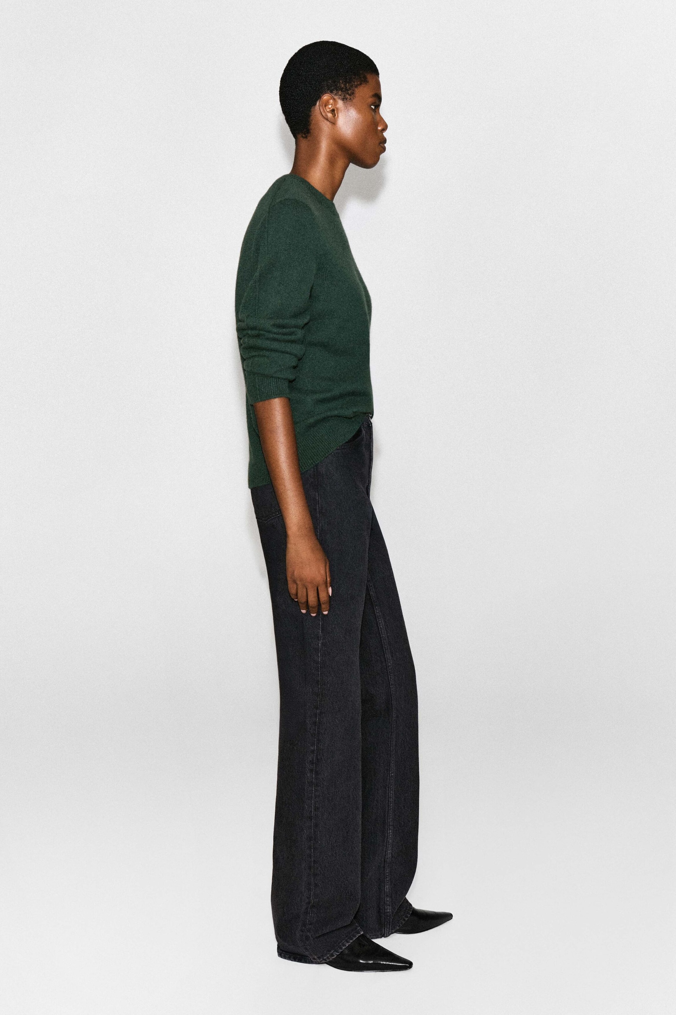 The model is wearing a dark green cashmere crewneck sweater while standing in profile. The long-sleeve sweater features ribbed trims and is styled with black wide-leg denim jeans. The model is wearing a dark green cashmere crewneck sweater while standing in profile. The long-sleeve sweater features ribbed trims and is styled with black wide-leg denim jeans.