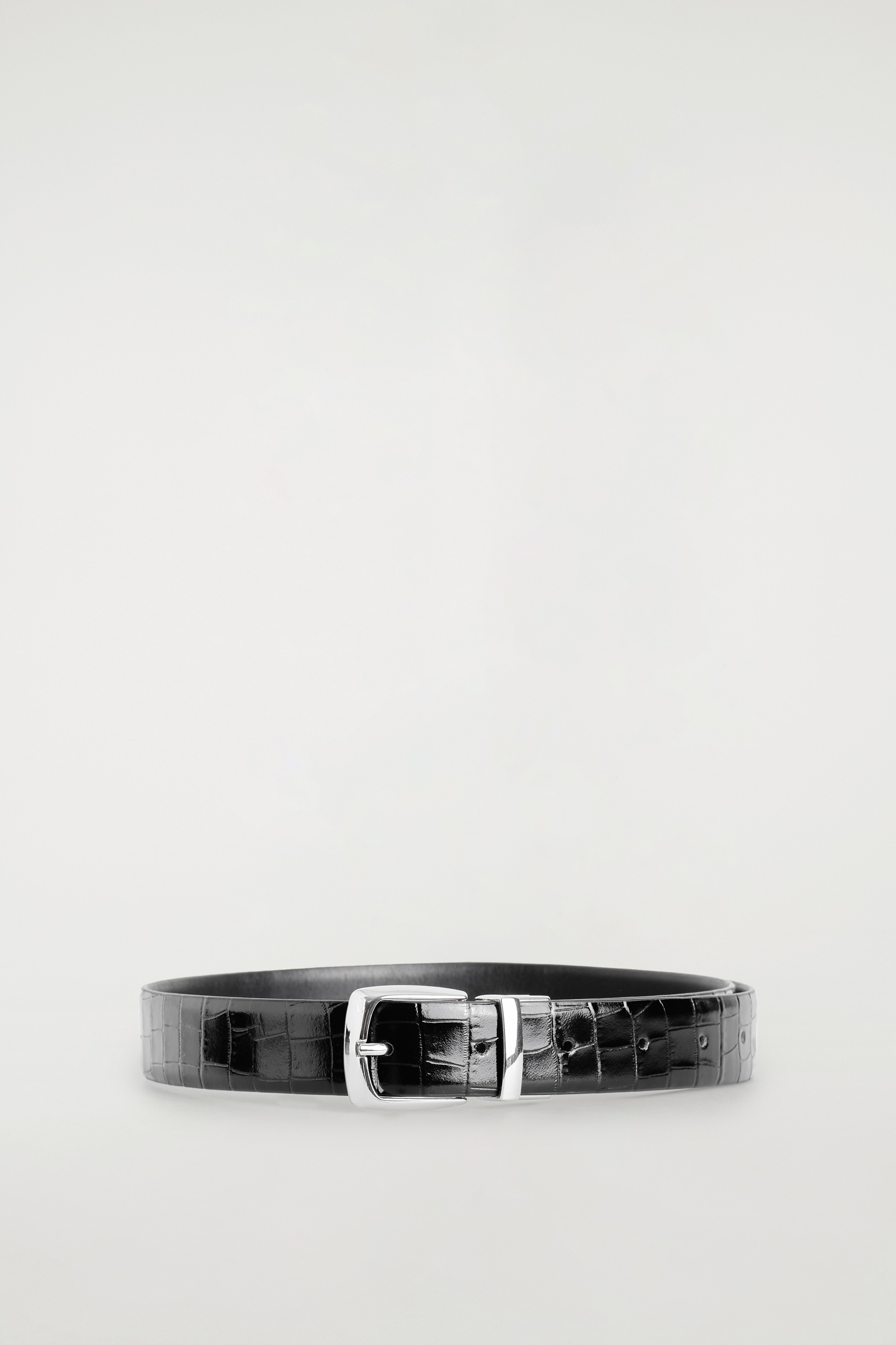 Een zwarte riem van leer met crocoprint en een zilverkleurige gesp wordt gedragen rond de taille van een zwarte leren rok. De riem is gestyled bij het model, dat ook een zwarte top en een open zwarte leren jas draagt.