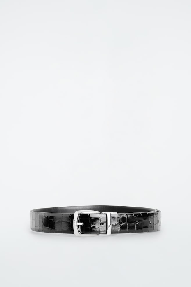 Une ceinture en cuir noir présentant une texture brillante embossée effet crocodile repose en boucle fermée, orientée vers lâavant. Elle met en valeur une boucle rectangulaire argentée au fini poli et un passant métallique assorti. - 1
