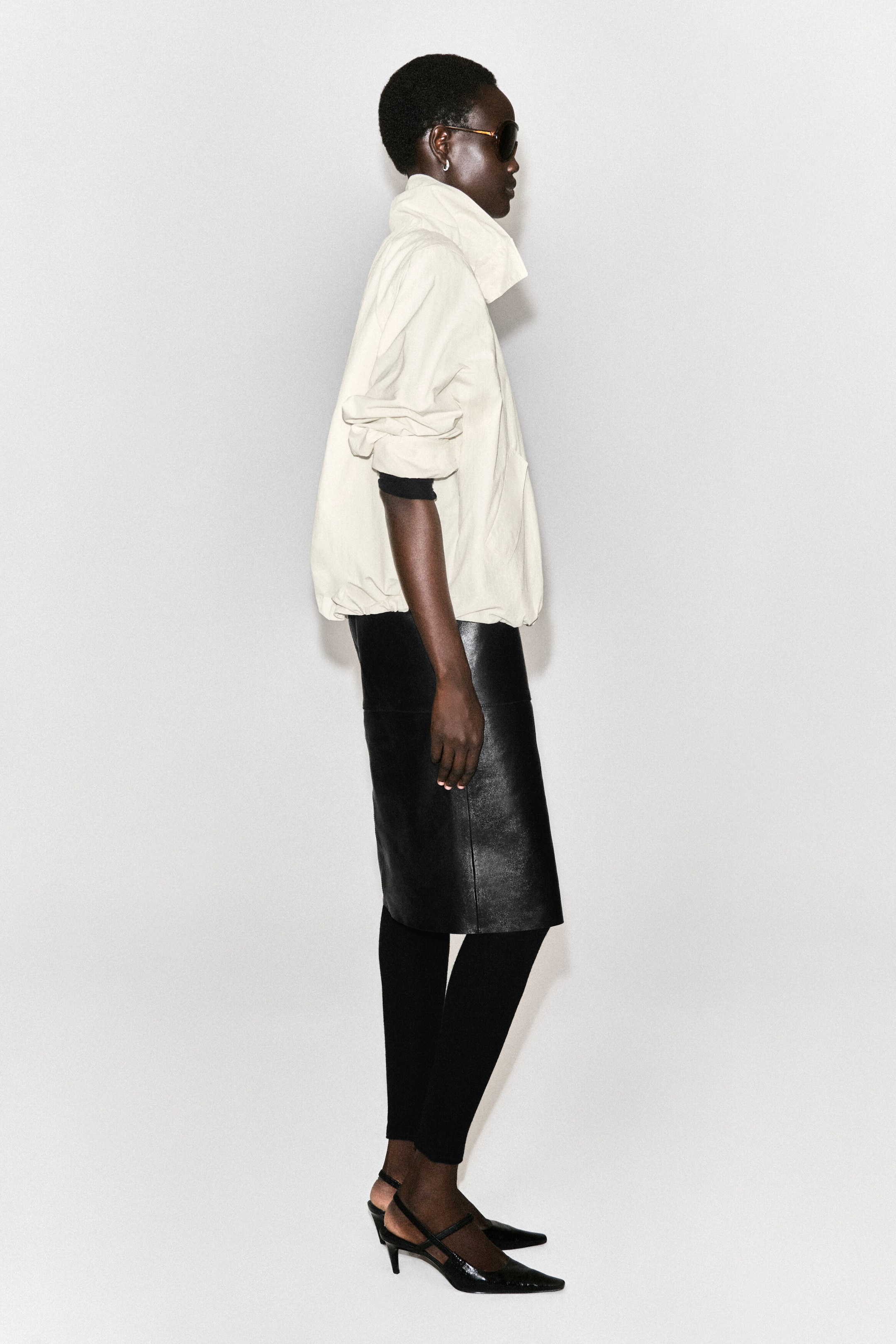 La veste en nylon blanc cassé à col cheminée est orientée vers la droite en vue de profil, mettant en valeur sa silhouette ample et décontractée avec un col montant et un ourlet froncé. Ses manches retroussées révèlent une sous-couche noire à manches longues au niveau de l’avant-bras. La veste en nylon blanc cassé à col cheminée est orientée vers la droite en vue de profil, mettant en valeur sa silhouette ample et décontractée avec un col montant et un ourlet froncé. Ses manches retroussées révèlent une sous-couche noire à manches longues au niveau de l’avant-bras.