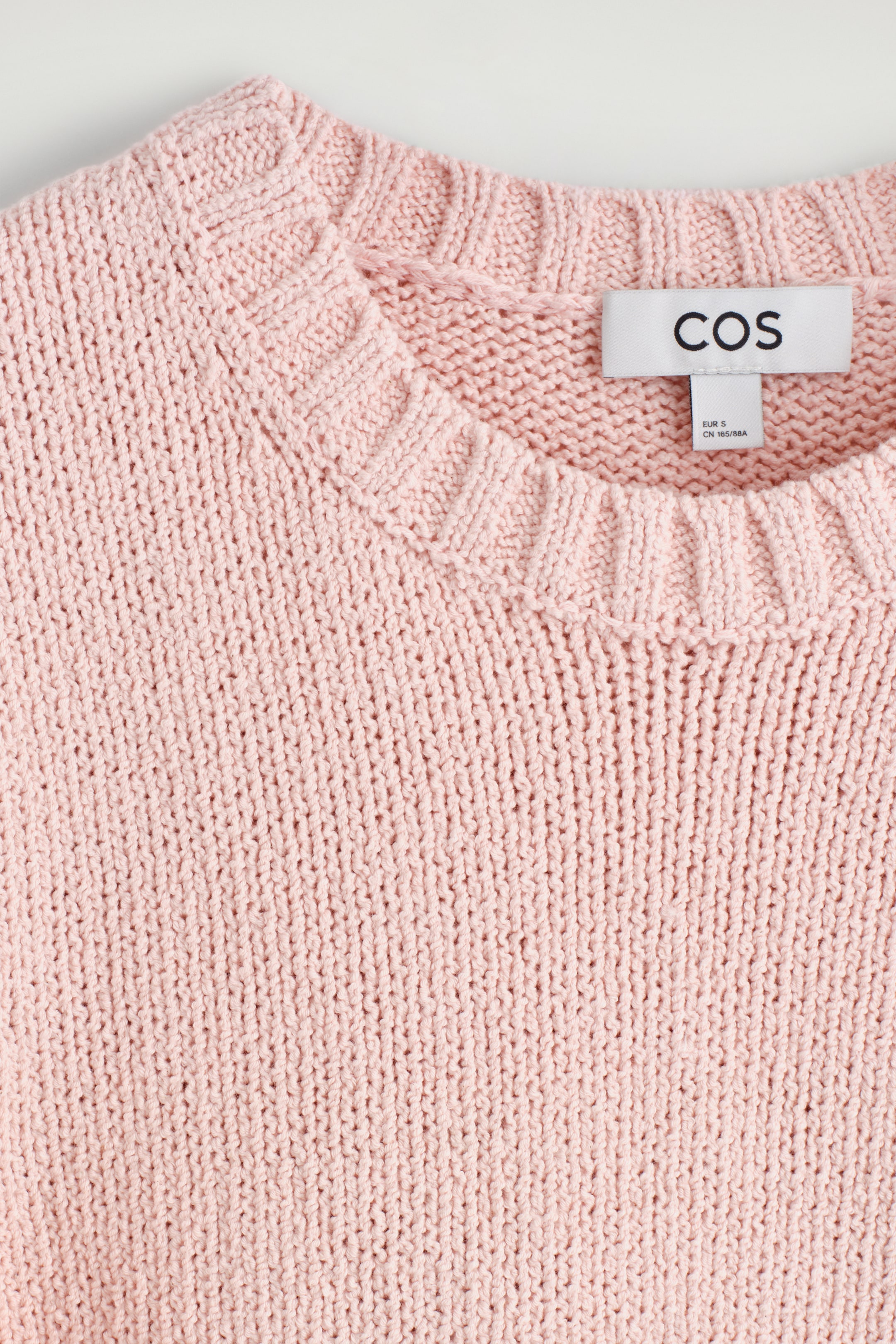 Größeres Bild anzeigen: Ein rosa gestricktes Kleidungsstück zeigt eine Nahaufnahme seines oberen Abschnitts, mit einem gerippten Rundhalsausschnitt und einem glatt rechts gestrickten Körper. Ein weißes Markenschild mit schwarzem Text ist auf der Innenseite des Kragens sichtbar.