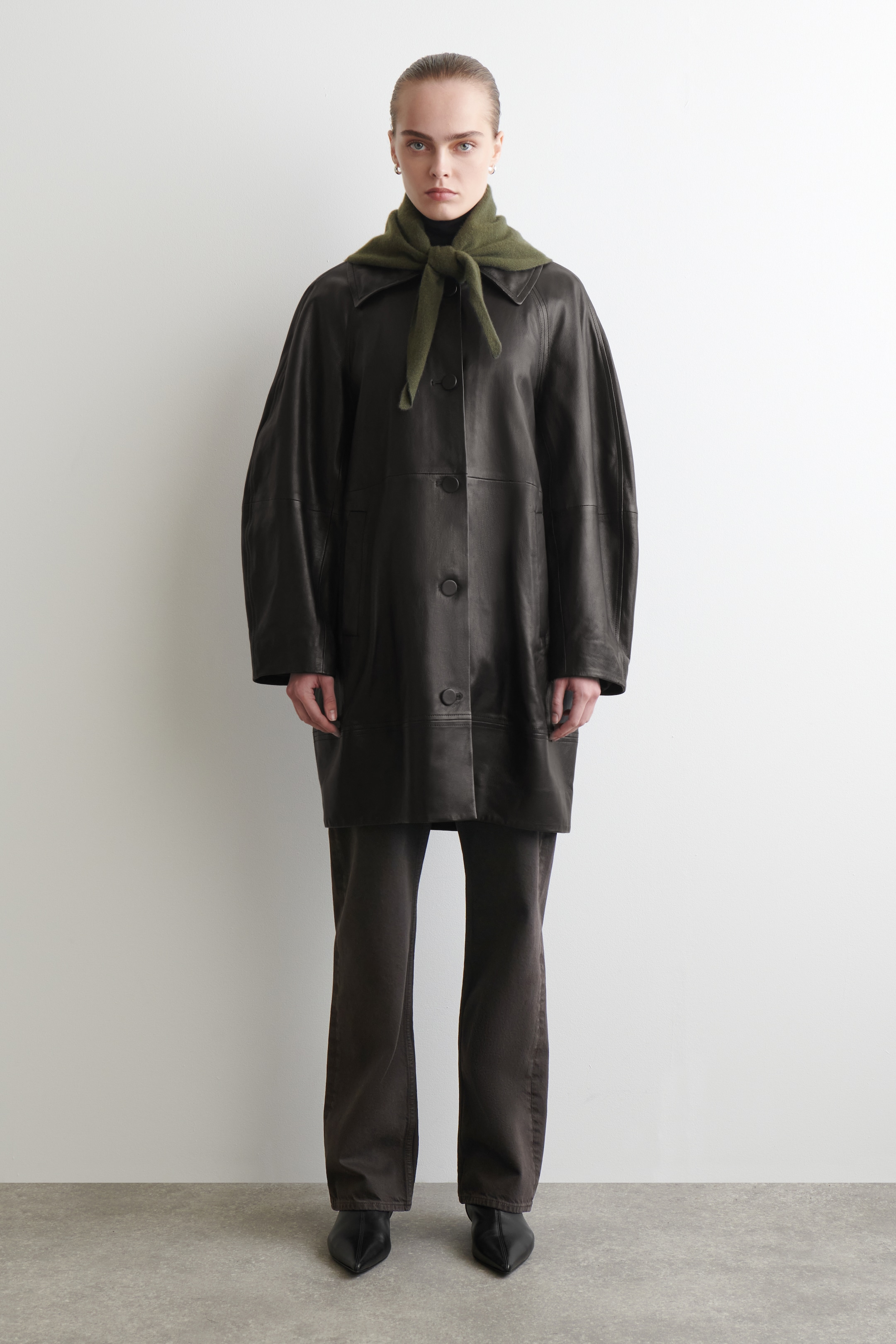 Visualizza immagine più grande: Un cappotto in pelle marrone scuro è rivolto in avanti, con un colletto aperto, una chiusura frontale con bottoni scuri e tasche a filetto verticali. Una sciarpa lavorata a maglia verde oliva riposa sulle spalle, formando un nodo sul davanti, mentre un dolcevita nero appare sotto il colletto del cappotto. Il cappotto presenta una silhouette ad A e le sue maniche lunghe mostrano una vestibilità ampia e rilassata.