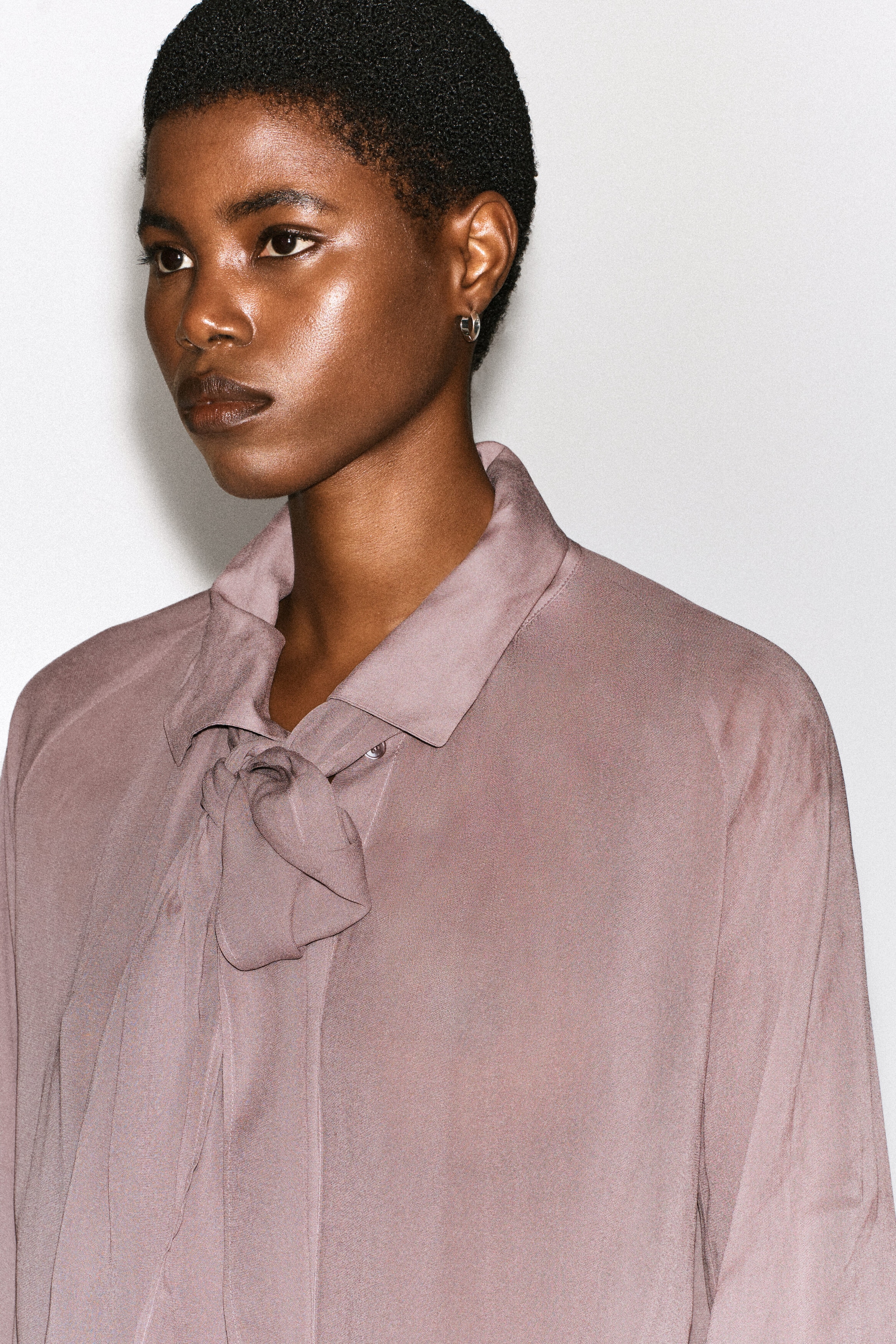 Le mannequin porte un haut fluide mauve avec détail foulard, doté d’un col classique et d’un détail foulard intégré noué lâchement à l’encolure.