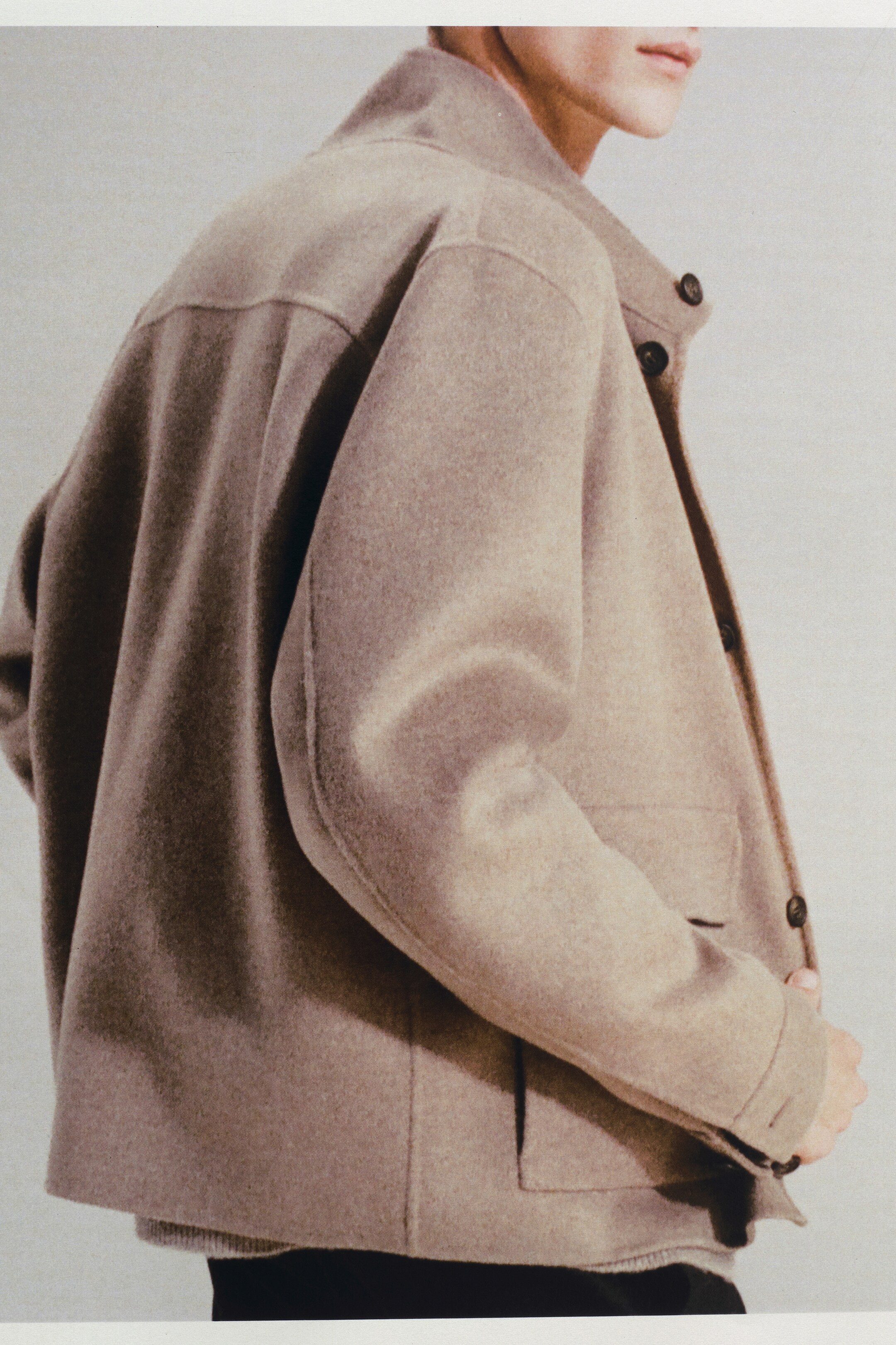 Größeres Bild anzeigen: Eine hell taupefarbene Wolljacke wird von einem Modell getragen, das leicht nach rechts geneigt ist und die Vorder- und Seite der Jacke präsentiert. Die Jacke verfügt über einen hohen Stehkragen mit sichtbaren Knöpfen, eine geknöpfte Frontleiste und zwei große aufgesetzte Taschen. Die Ärmel erscheinen leicht gebogen, und die Manschetten sind geknöpft. Ein heller gefärbter Pullover ist unter der Jacke sichtbar.