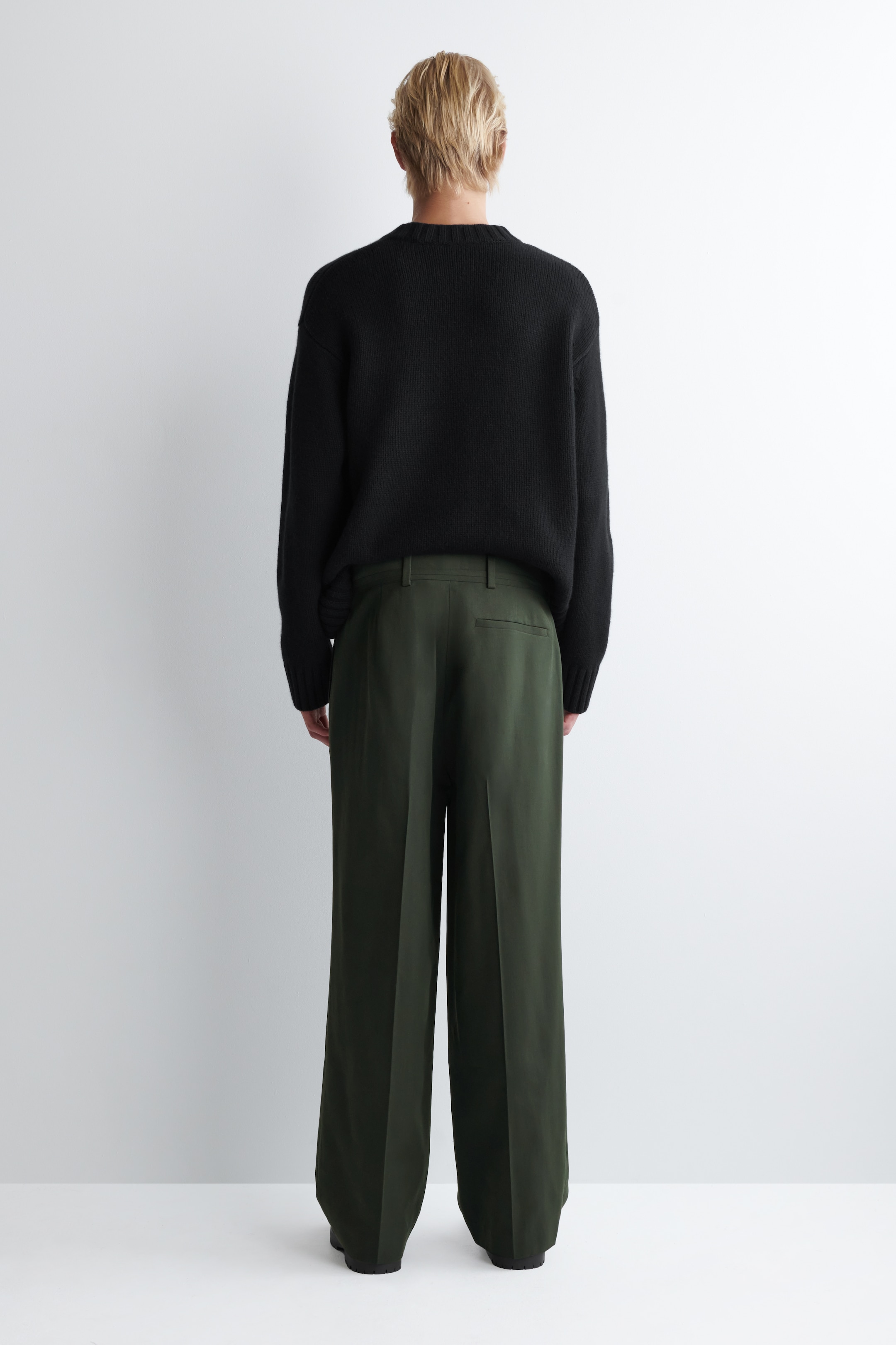 Ingrandisci l'immagine: Un maglione nero a girocollo con vestibilità rilassata è indossato, visto da dietro, con maniche lunghe. È abbinato a pantaloni verde oliva scuro a gamba larga che mostrano passanti per cintura, una tasca a filetto singola sul lato destro del retro e una piega centrale prominente lungo ciascuna gamba, che si accumula all'orlo.