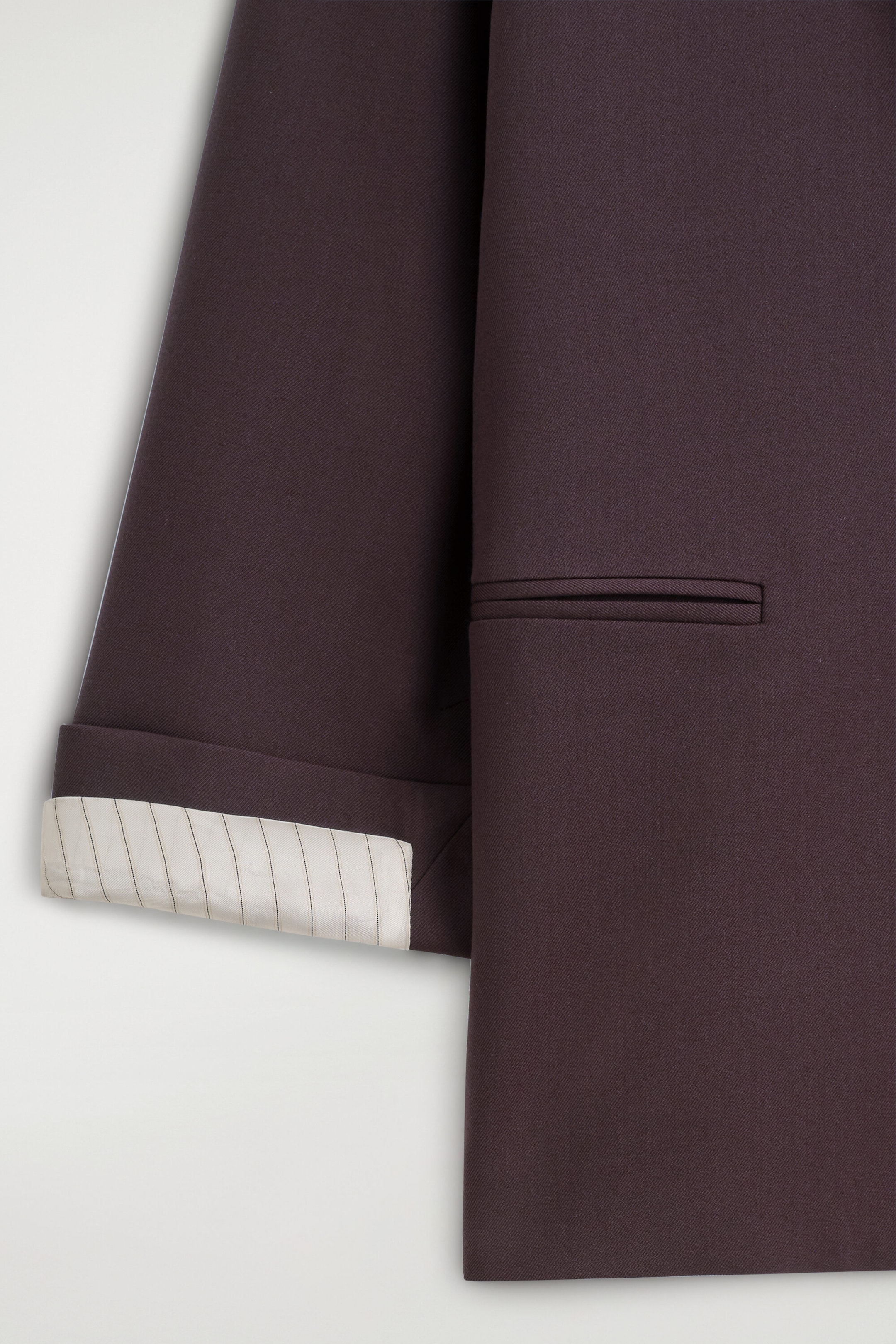 Eine Nahaufnahme eines dunkel pflaumenfarbenen Blazers zeigt die Vorderseite und einen Ärmel. Der Ärmel hat einen hochgekrempelten Umschlag, der ein cremefarbenes, feingestreiftes Futter freilegt, während der Körper eine horizontale Doppelpaspeltasche aufweist. Eine Nahaufnahme eines dunkel pflaumenfarbenen Blazers zeigt die Vorderseite und einen Ärmel. Der Ärmel hat einen hochgekrempelten Umschlag, der ein cremefarbenes, feingestreiftes Futter freilegt, während der Körper eine horizontale Doppelpaspeltasche aufweist.