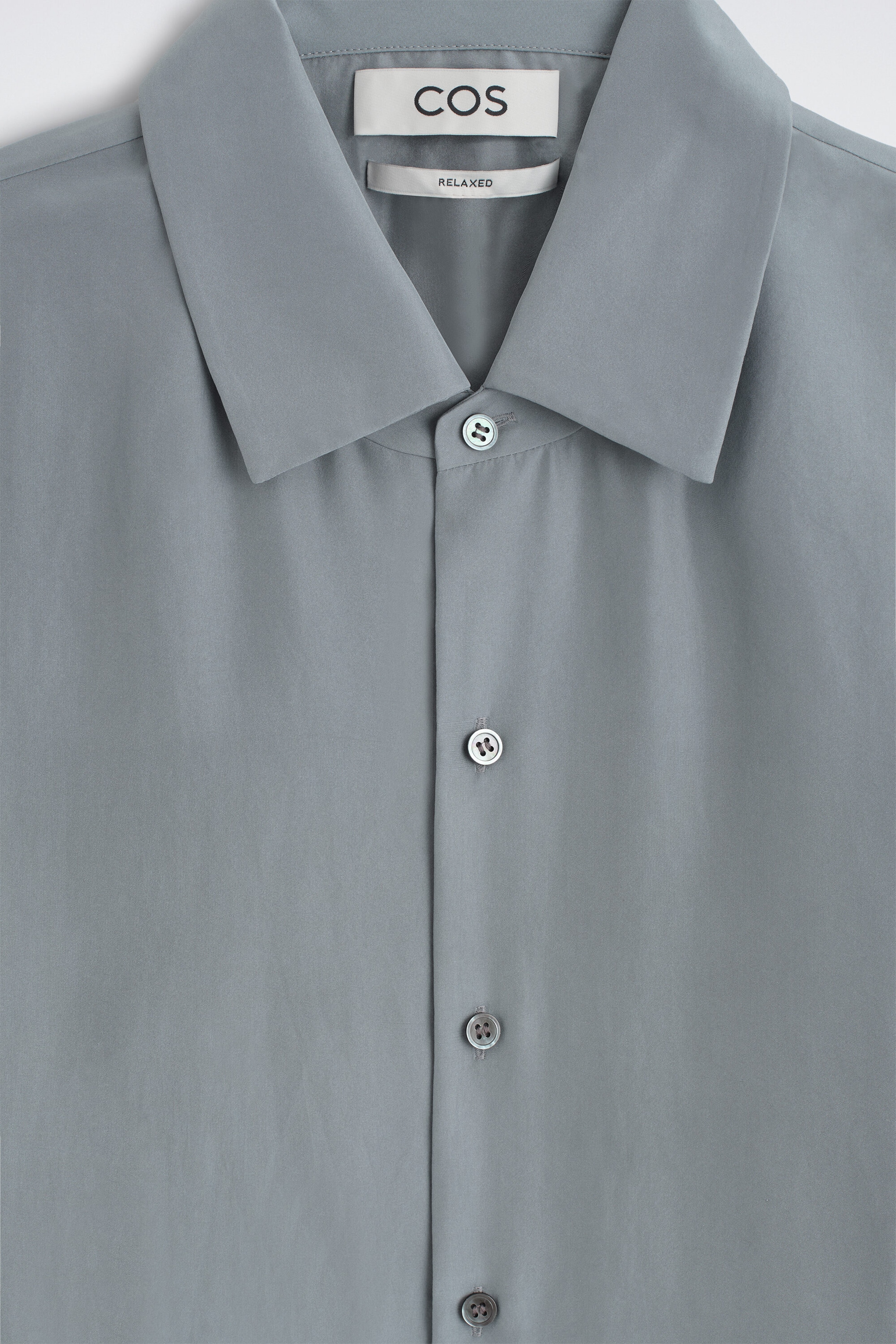 A close-up front view shows a relaxed silk shirt in a muted slate grey colour, highlighting its pointed collar and button-down front placket. The smooth silk fabric has a soft matte finish, with four matching round buttons visible along the centre seam. A close-up front view shows a relaxed silk shirt in a muted slate grey colour, highlighting its pointed collar and button-down front placket. The smooth silk fabric has a soft matte finish, with four matching round buttons visible along the centre seam.