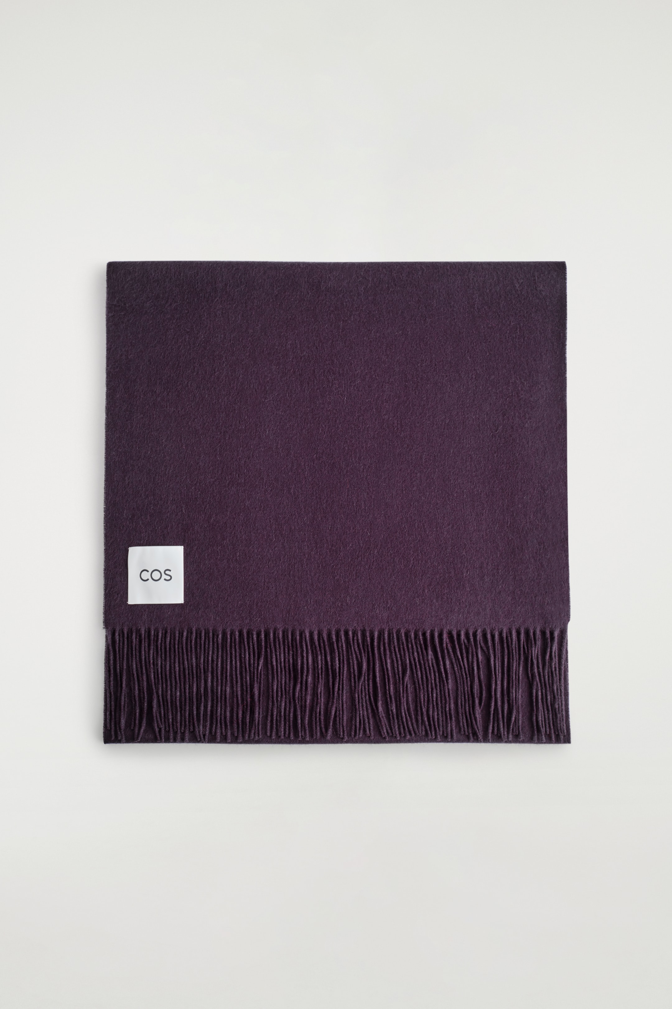 Agrandir l'image: Une large écharpe de ton bordeaux est présentée vue de dessus, avec une texture lisse et des franges le long de son bord inférieur. Une étiquette blanche COS est visible en haut à gauche de l'écharpe.