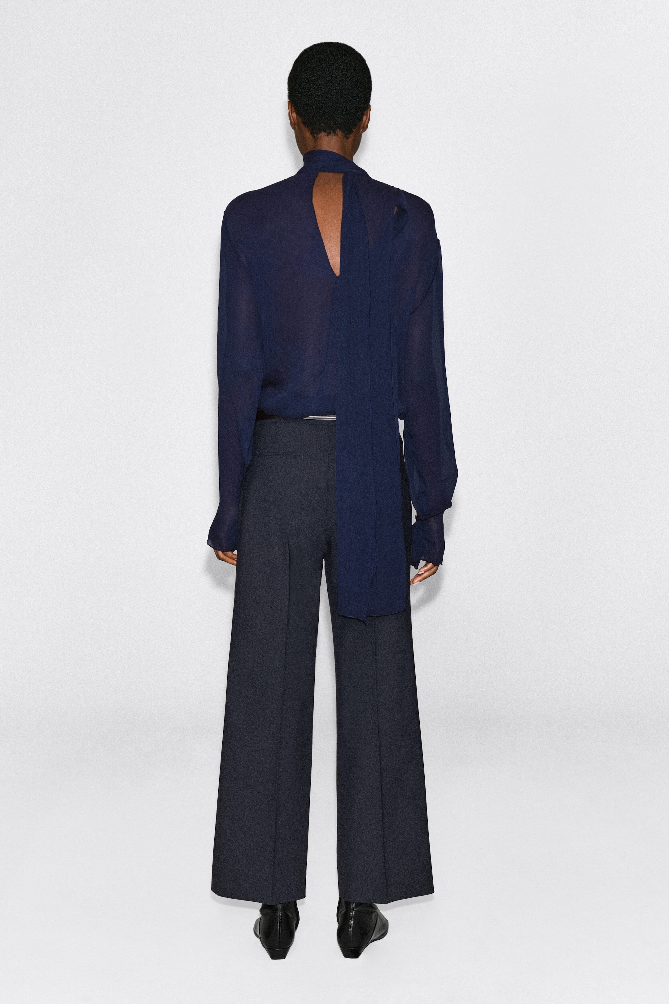 A semi-sheer, dark blue long-sleeved blouse is being worn by the model, shown from the back. The top features a high neck with a long, draped scarf hanging down the back, a keyhole cut-out, and is tucked into dark, wide-leg trousers. A semi-sheer, dark blue long-sleeved blouse is being worn by the model, shown from the back. The top features a high neck with a long, draped scarf hanging down the back, a keyhole cut-out, and is tucked into dark, wide-leg trousers.