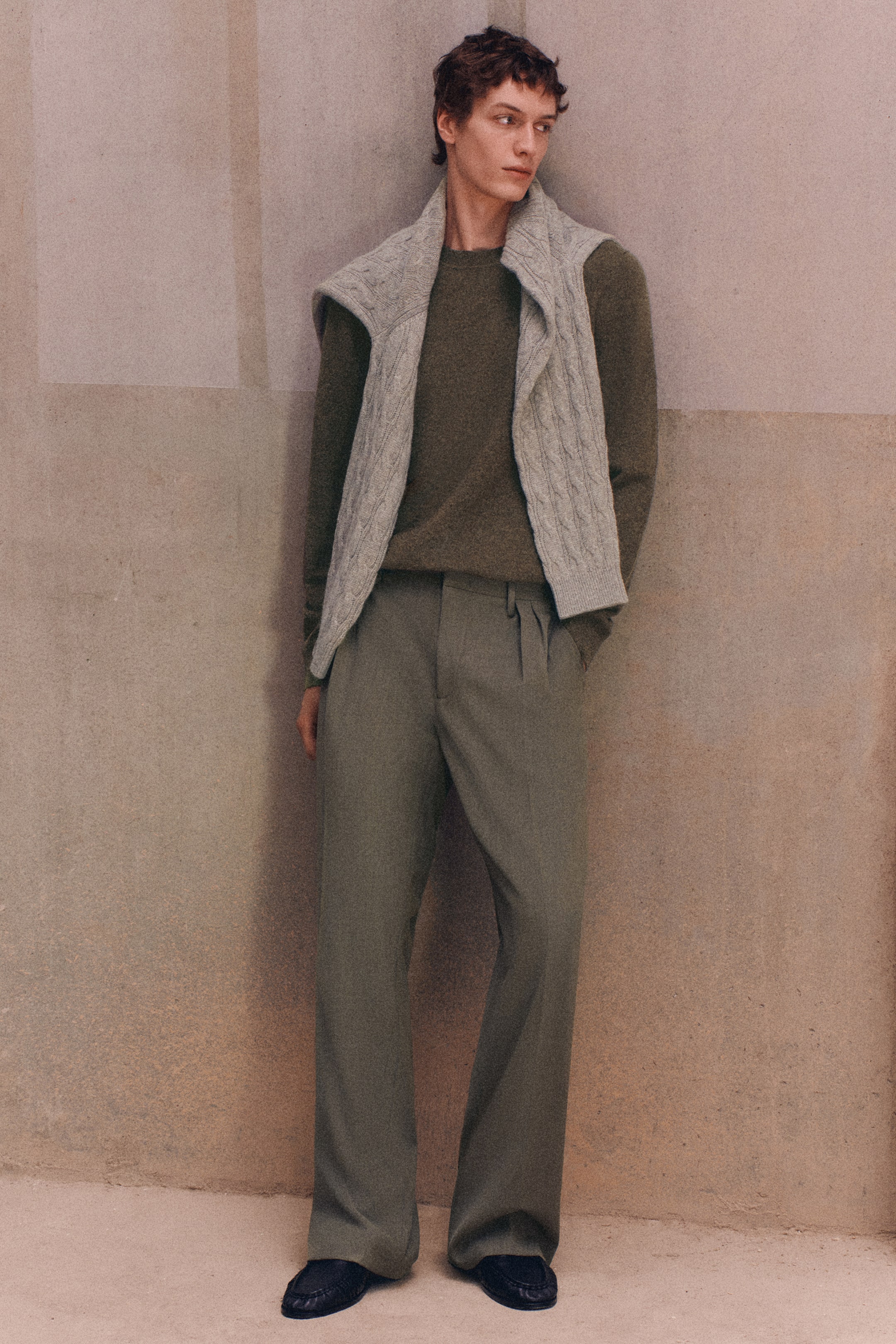Visualizza l'immagine ingrandita: Il pullover girocollo in cashmere verde oliva e i pantaloni a gamba larga in misto lana, dal taglio rilassato, verde salvia, sono mostrati frontalmente su una modella. Il pullover a maniche lunghe è abbinato a un maglione a trecce grigio chiaro drappeggiato sulle spalle, mentre i pantaloni presentano pieghe frontali e cadono in una silhouette rilassata a gamba larga.