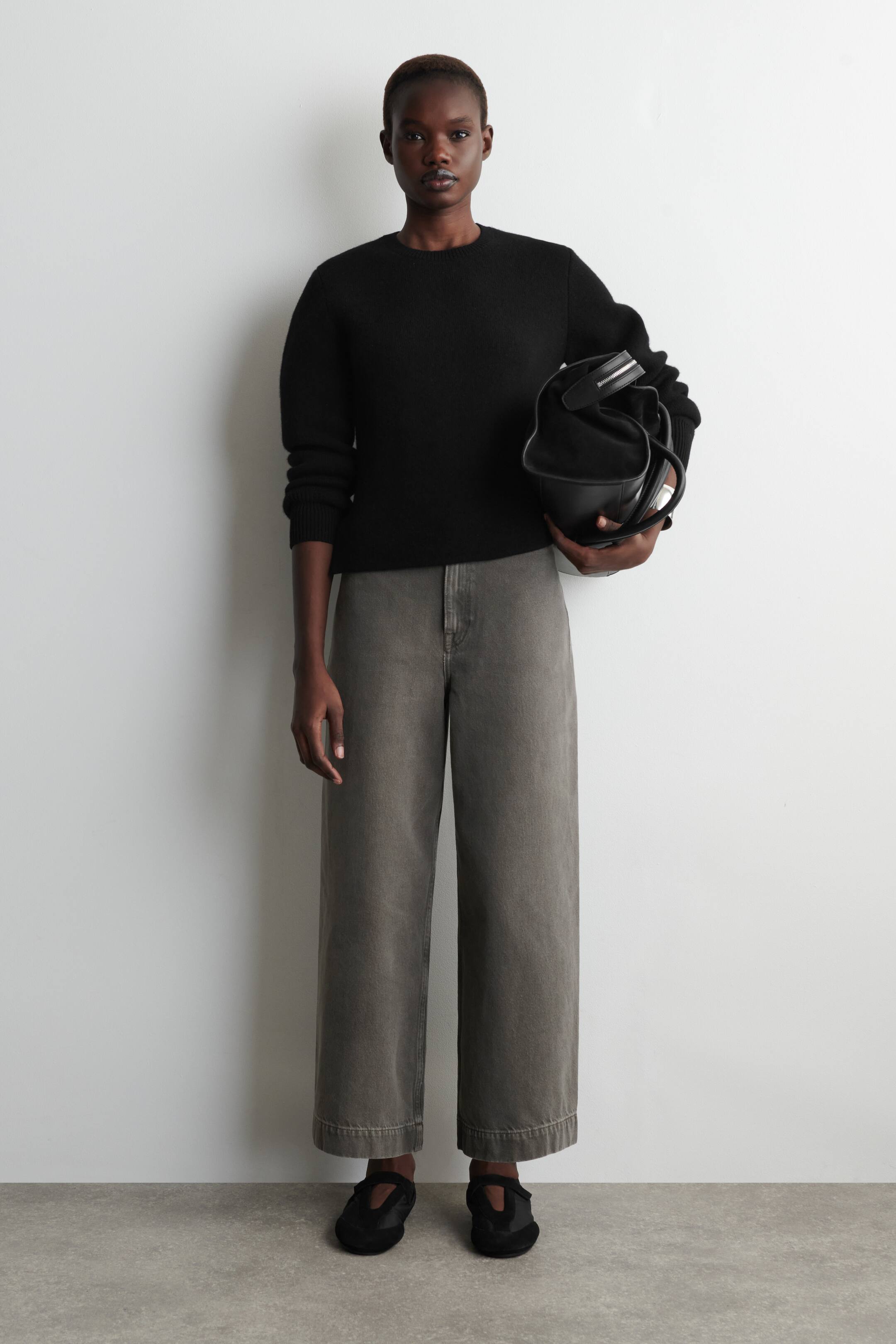 Größeres Bild anzeigen: Eine Person steht und trägt einen schwarzen Rundhalspullover mit gerippten Bündchen, kombiniert mit grauen, weit geschnittenen Hosen mit verkürztem Saum. Dazu trägt sie schwarze Slipper mit einem Riemen über dem Spann.