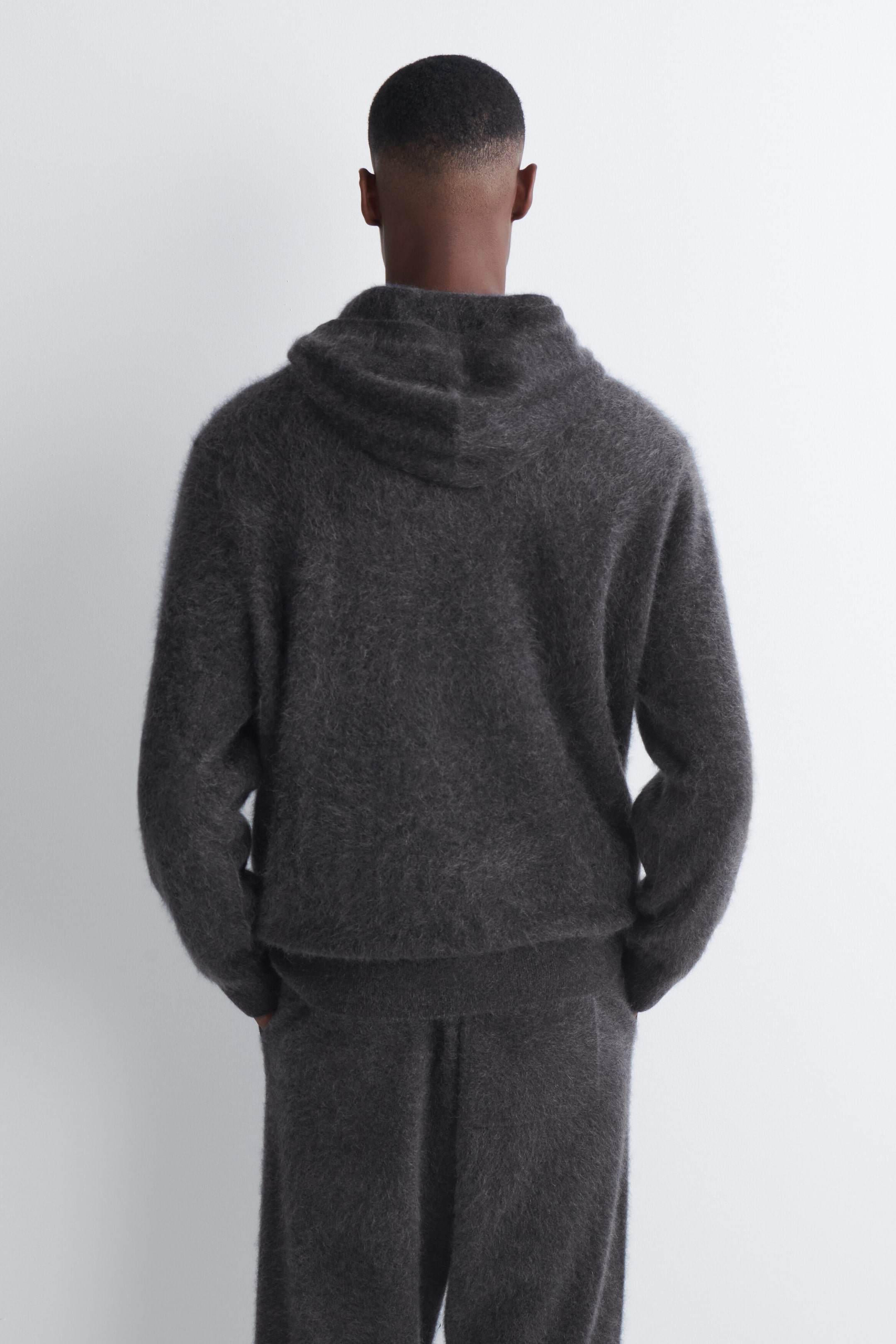 A dark gray brushed cashmere hoodie with a soft, fuzzy texture is worn by the model and shown from the back. The hoodie features an attached hood and a ribbed hem, and is paired with matching knit trousers.