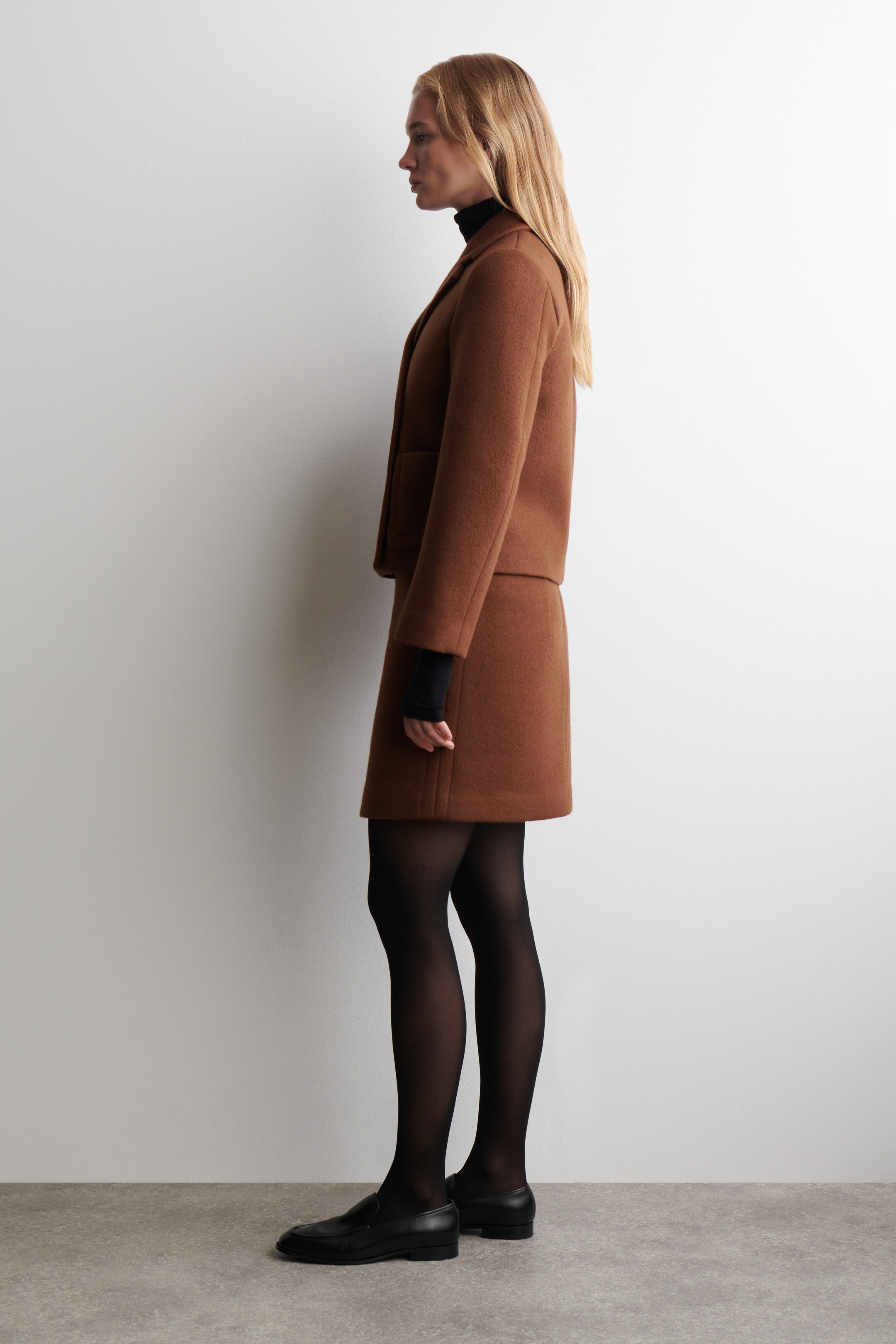 View larger image: A brown jacket and skirt set is being displayed from a side view, featuring a jacket with notch lapels and a patch pocket, and a matching A-line skirt falling just above the knee. A black turtleneck is layering underneath the jacket.
