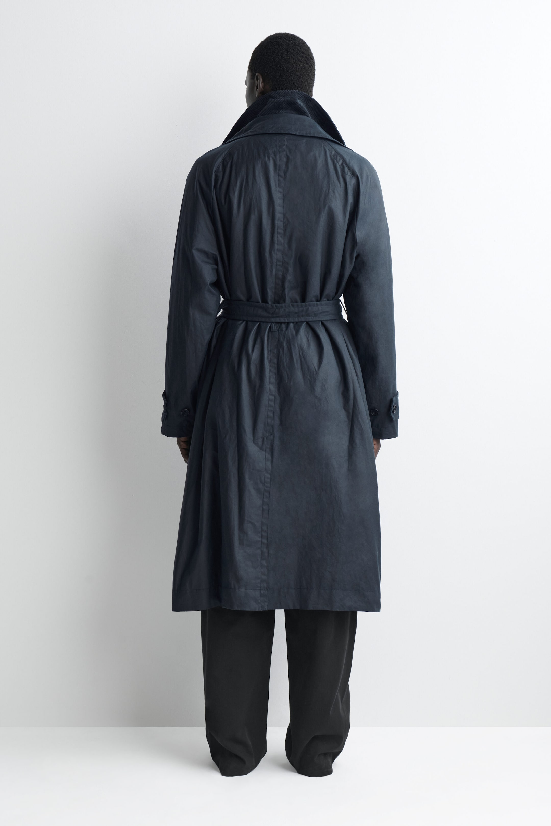 This dark navy trench coat is shown from the back, featuring a relaxed fit cinched at the waist with a matching fabric belt. The coat features a high, turned-up funnel collar with a textured corduroy lining and long sleeves with button-tab cuffs, falling to a mid-calf length over black trousers. This dark navy trench coat is shown from the back, featuring a relaxed fit cinched at the waist with a matching fabric belt. The coat features a high, turned-up funnel collar with a textured corduroy lining and long sleeves with button-tab cuffs, falling to a mid-calf length over black trousers.