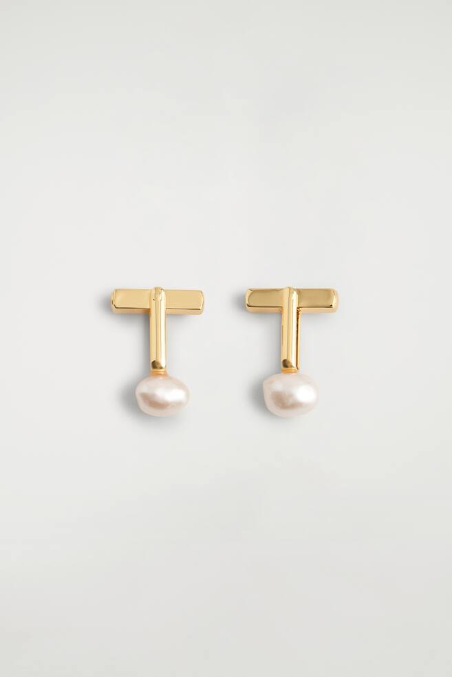 A pair of gold-toned cufflinks is shown front-on, featuring a polished T-bar design connected by a straight post to an irregular white baroque pearl. - 1