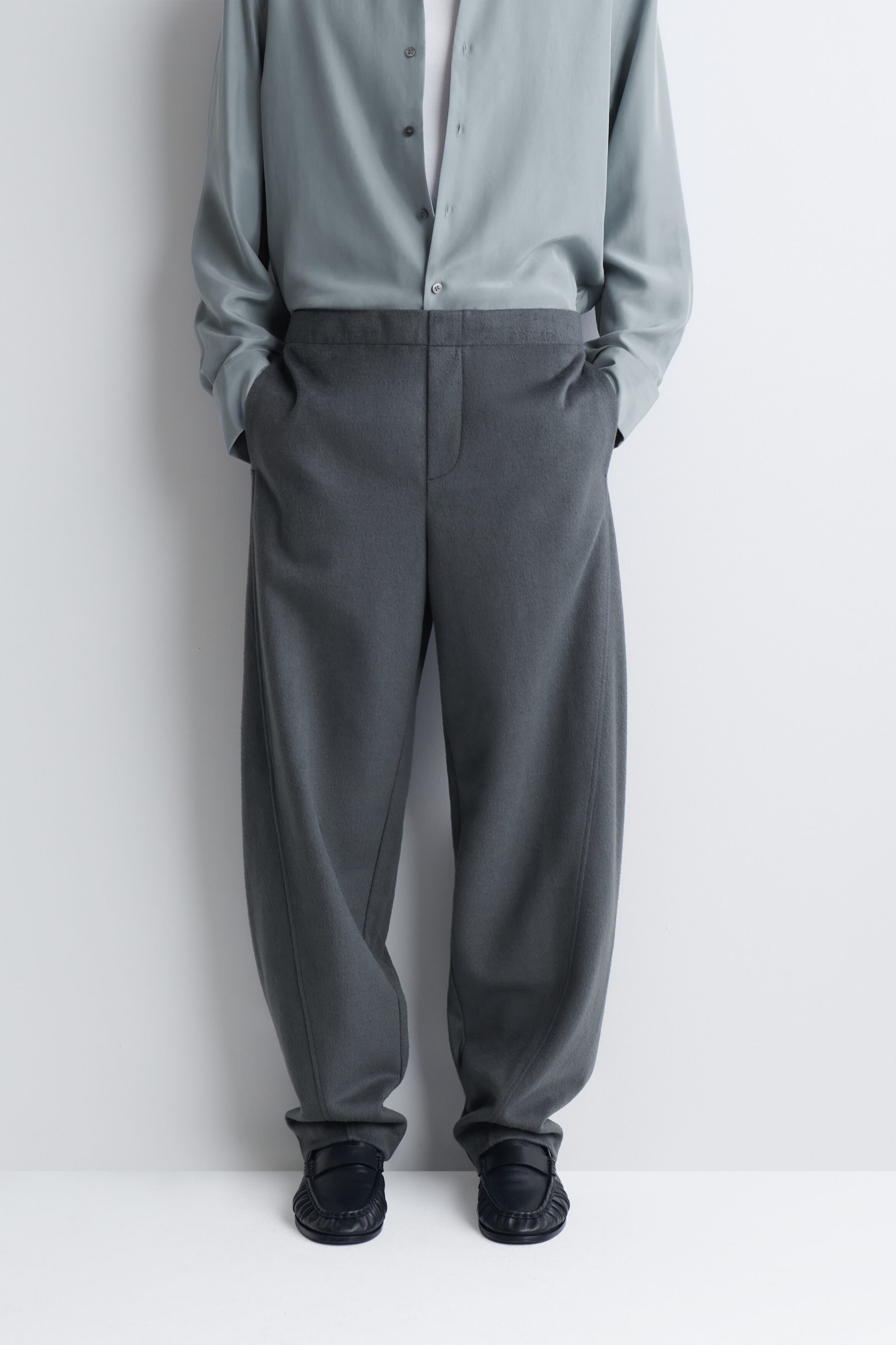 Le mannequin porte un pantalon large gris foncé, les mains glissées dans les poches latérales. Le pantalon présente une silhouette décontractée légèrement arrondie et est agencé avec une chemise à manches longues gris clair rentrée dans le pantalon, ainsi qu’avec des chaussures en cuir noir.