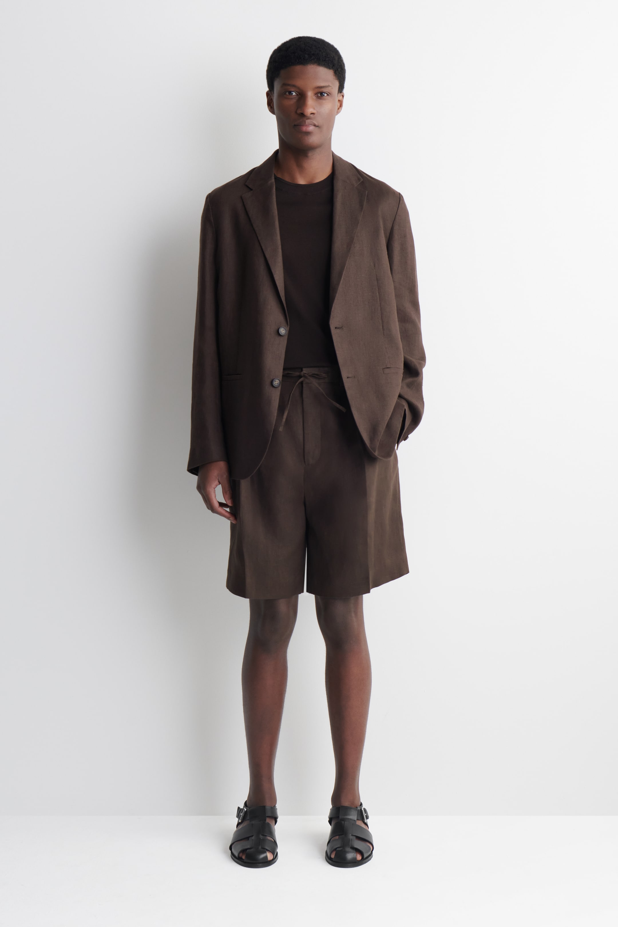 Ver imagem maior: Um blazer castanho para homem com lapelas em bico e dois botões está a ser usado aberto sobre uma t-shirt de gola redonda castanho-escuro, combinado com calções castanhos de cordão a condizer. Sandálias pretas com várias tiras completam este conjunto de ajuste descontraído e virado para a frente.
