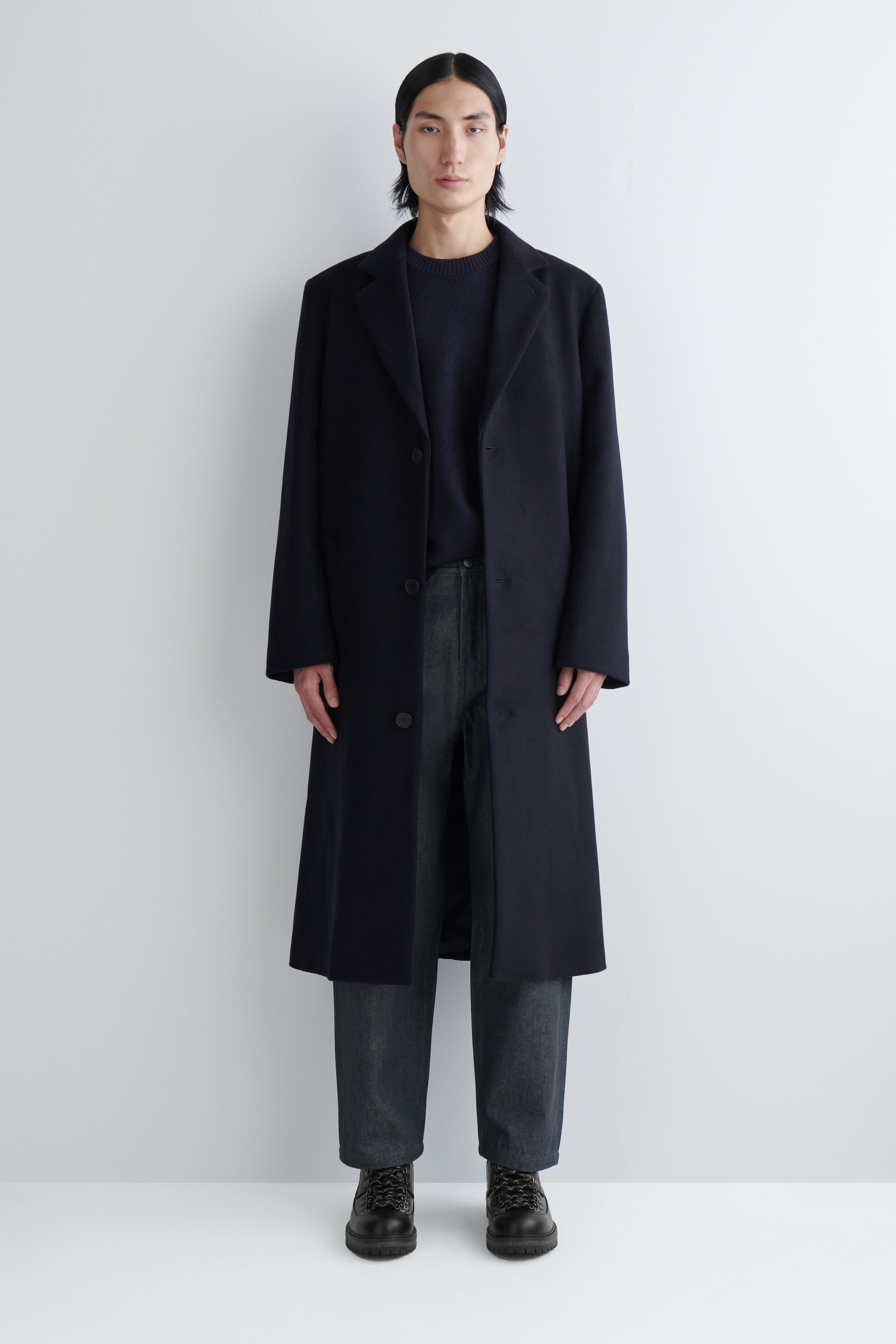 Agrandir l'image: Un long manteau bleu marine foncé est présenté de face, avec des revers crantés et une fermeture à bouton simple. Il s'étend sous le genou, avec un pull à col rond foncé et un pantalon foncé visibles en dessous, mettant en avant la coupe décontractée du manteau.