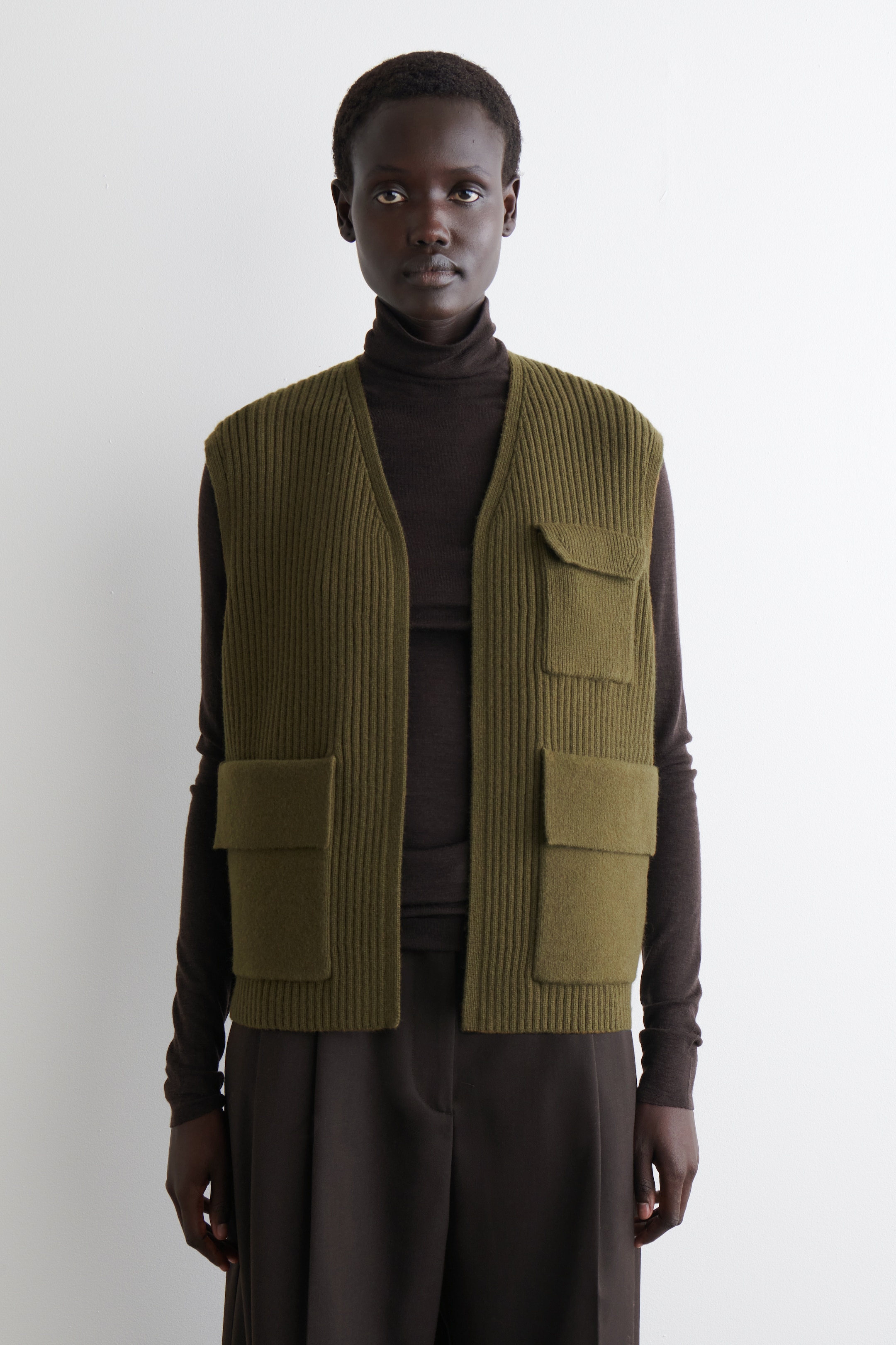 View larger image: An olive green ribbed knit vest is being worn, facing forward, showcasing its V-neck design. It features four prominent patch pockets with flaps, two positioned on the upper chest and two lower down, and is layered over a dark brown turtleneck.