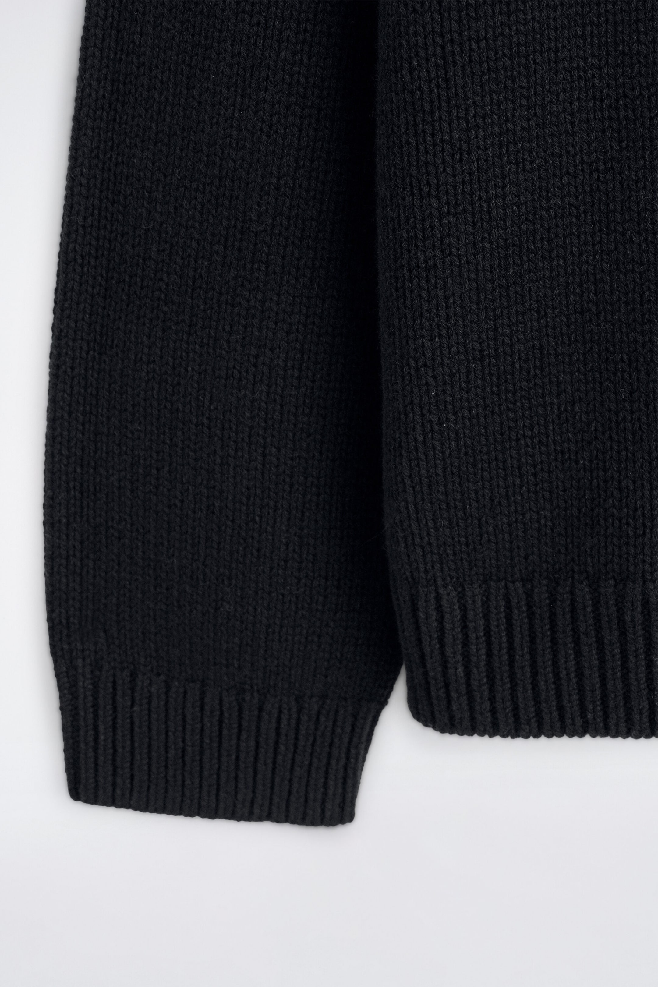 A close-up of a black chunky-knit jumper highlighting the textured ribbed cuff on the sleeve and the matching ribbed hem at the bottom. The garment displays a thick, vertical ribbed-knit structure. A close-up of a black chunky-knit jumper highlighting the textured ribbed cuff on the sleeve and the matching ribbed hem at the bottom. The garment displays a thick, vertical ribbed-knit structure.