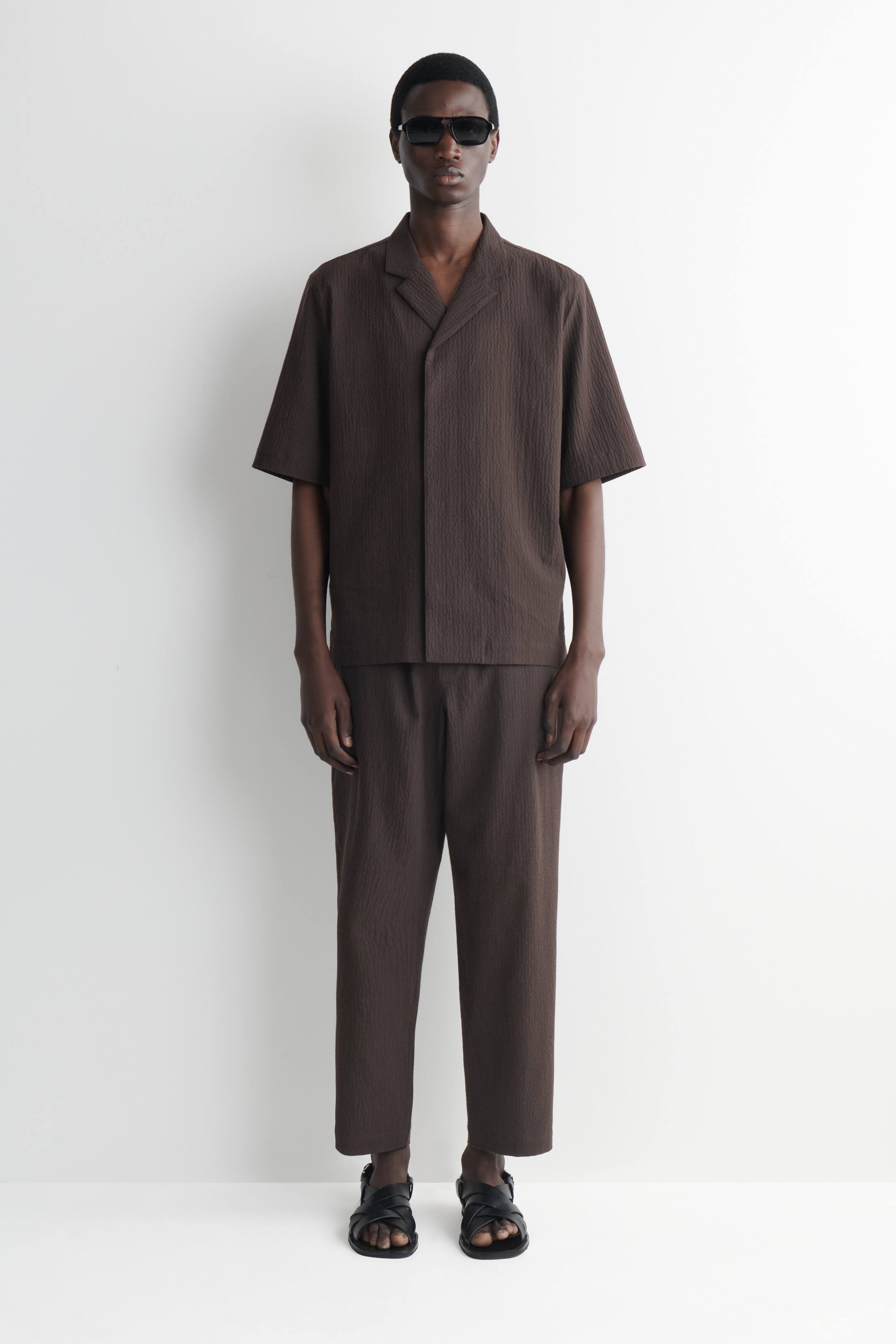 Visa större bild: Ett brunt tvådelat set presenteras, bestående av en kortärmad skjorta med reverkrage och matchande korta byxor. Båda plaggen är designade med ett subtilt texturerat tyg och en avslappnad passform, vända framåt.