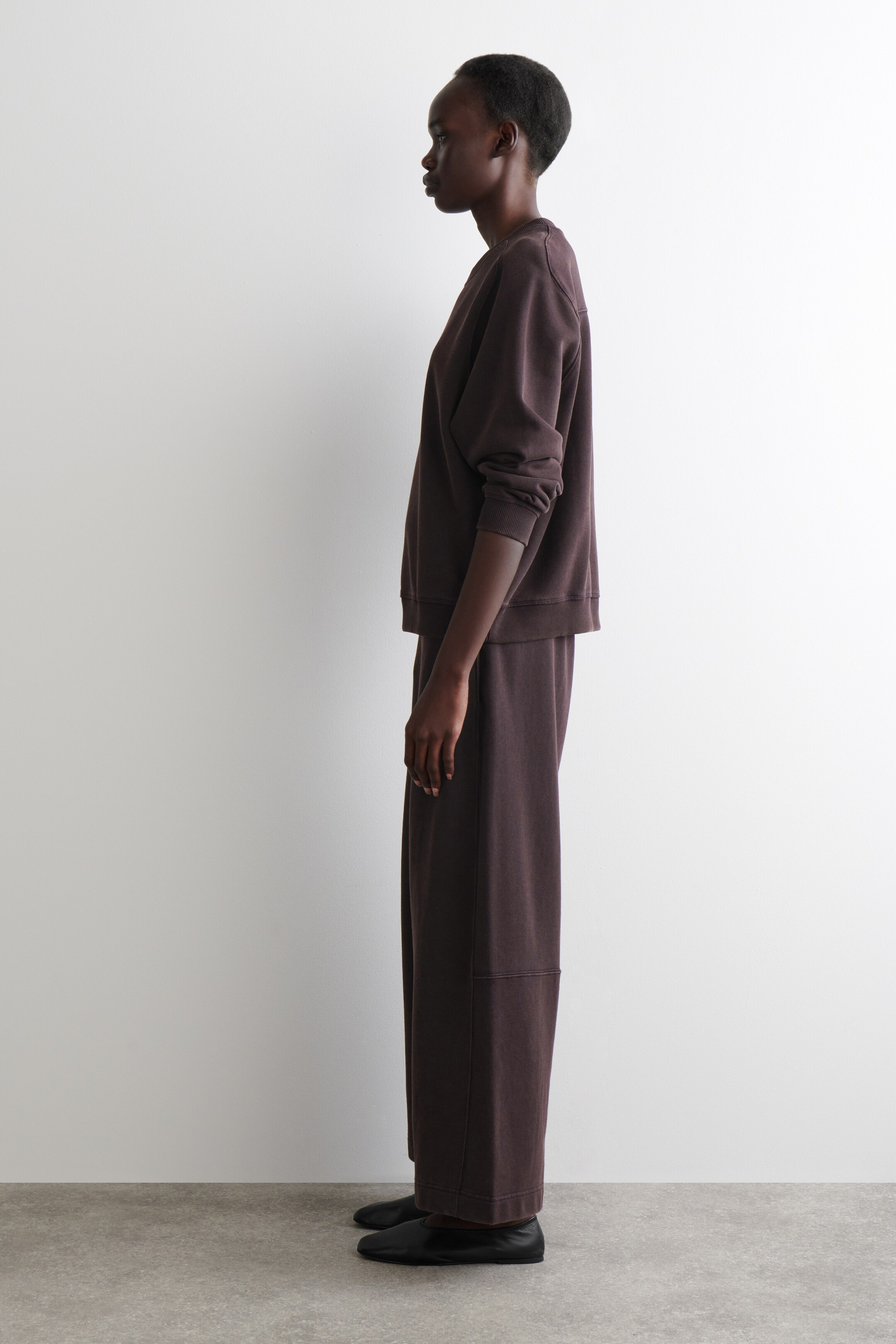 Visualizza immagine più grande: Un set da loungewear a due pezzi di colore marrone scuro viene presentato di profilo. La felpa con scollo rotondo a vestibilità rilassata presenta polsini e orlo a coste, mentre i pantaloni a gamba larga abbinati mostrano una cucitura orizzontale sulla parte inferiore della gamba, il tutto abbinato a scarpe nere piatte.