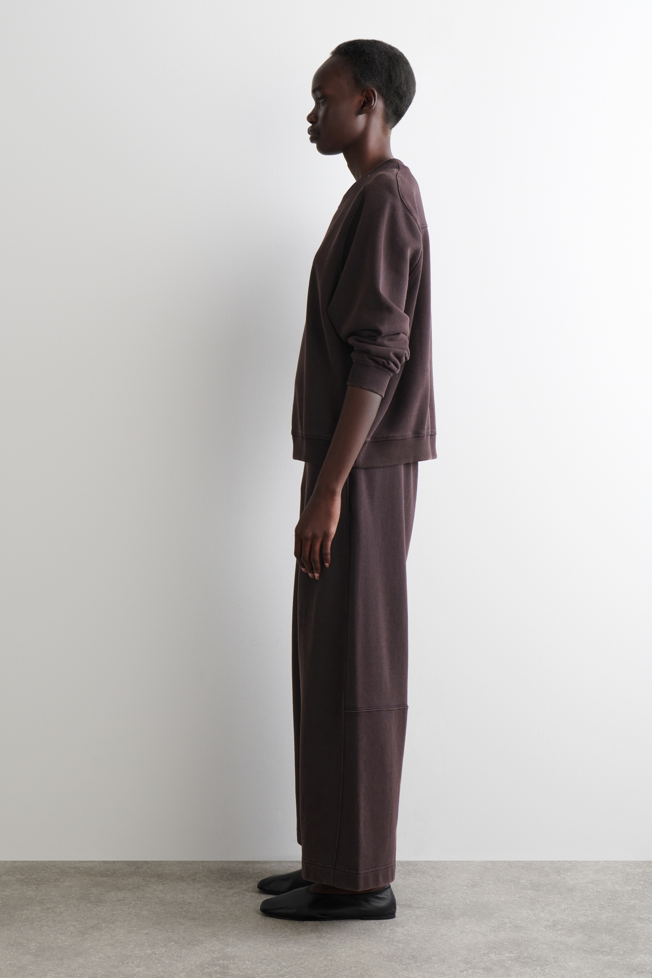Agrandir l'image: Un ensemble de détente deux pièces de couleur brun foncé est présenté de profil. Le sweat-shirt à col rond et coupe décontractée présente des poignets et un ourlet côtelés, tandis que le pantalon large assorti affiche une couture horizontale sur le bas de la jambe, le tout étant stylé avec des chaussures noires plates.