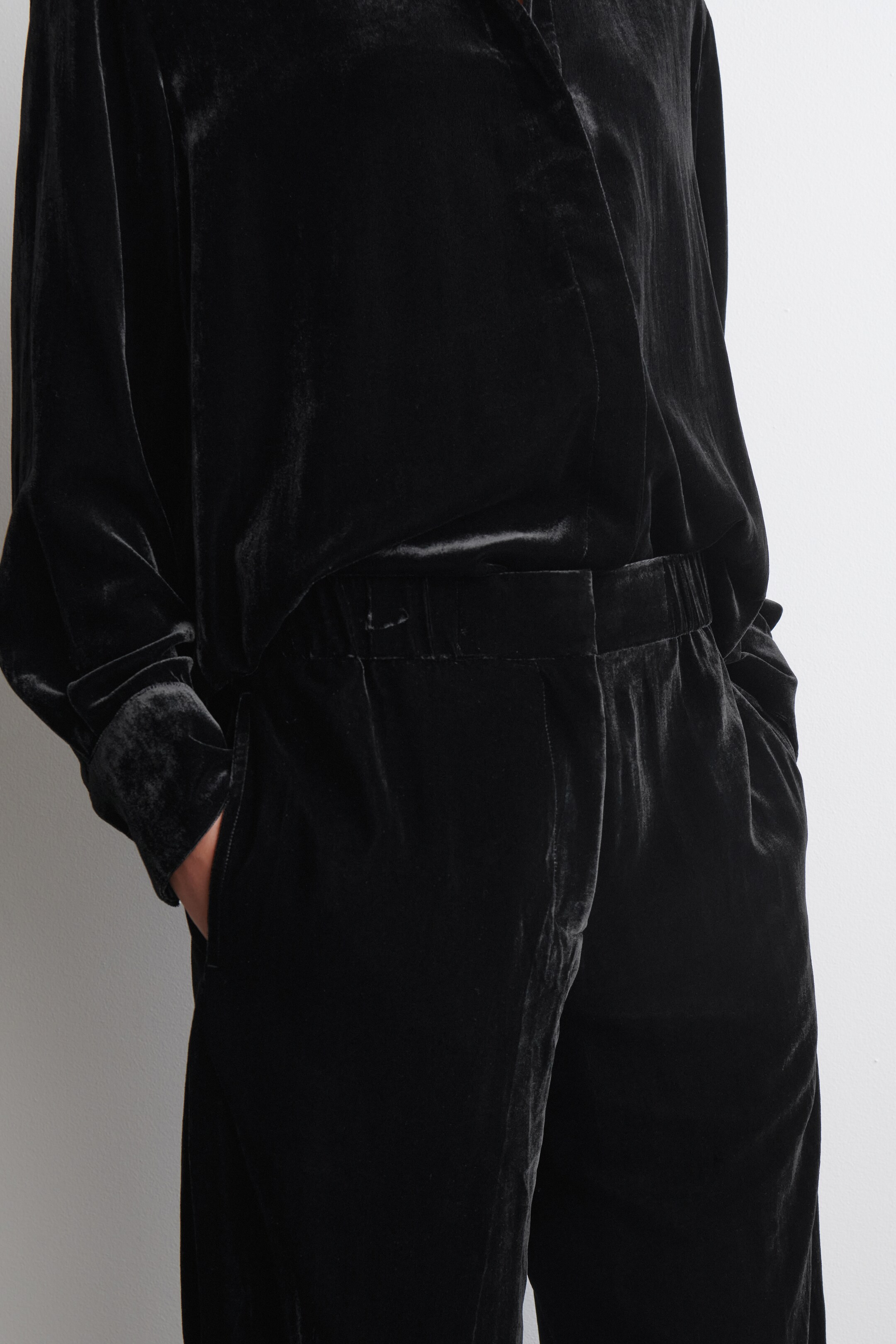 Agrandir l'image: Un ensemble chemise et pantalon en velours noir est présenté sous un léger angle latéral. La chemise à manches longues présente une coupe décontractée, une ouverture en V au niveau du cou, et un tissu drapé. Le pantalon en velours assorti met en avant une ceinture élastiquée et des poches latérales, avec une main reposant dans l'une d'elles.