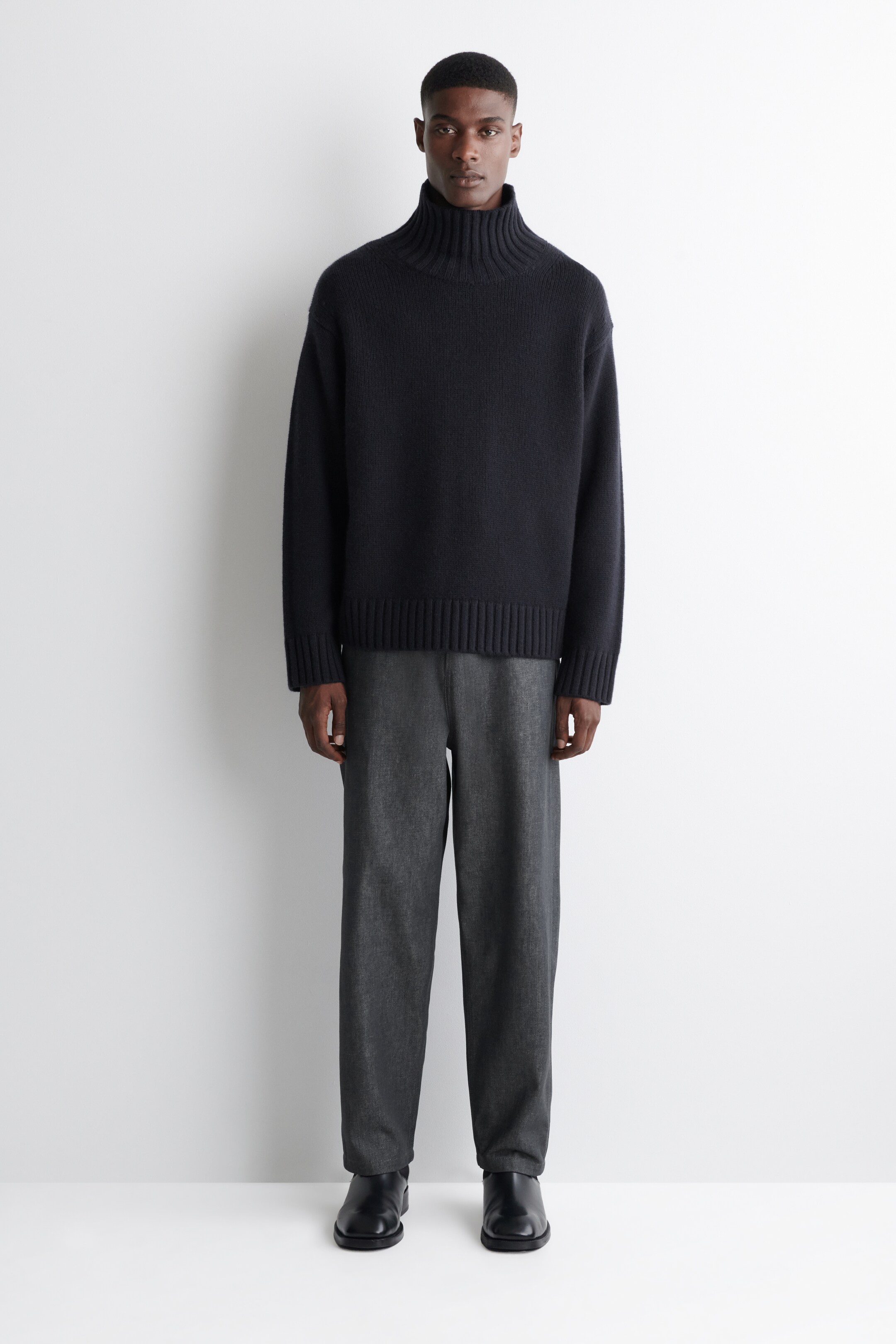 Agrandir l'image: Un pull à col roulé bleu foncé est porté, avec un col haut côtelé, des poignets côtelés et un ourlet côtelé. Il est assorti à un pantalon gris foncé à coupe décontractée et des bottes noires à bout carré, tous les articles étant orientés vers l'avant, mettant en valeur leur design et leur coupe.