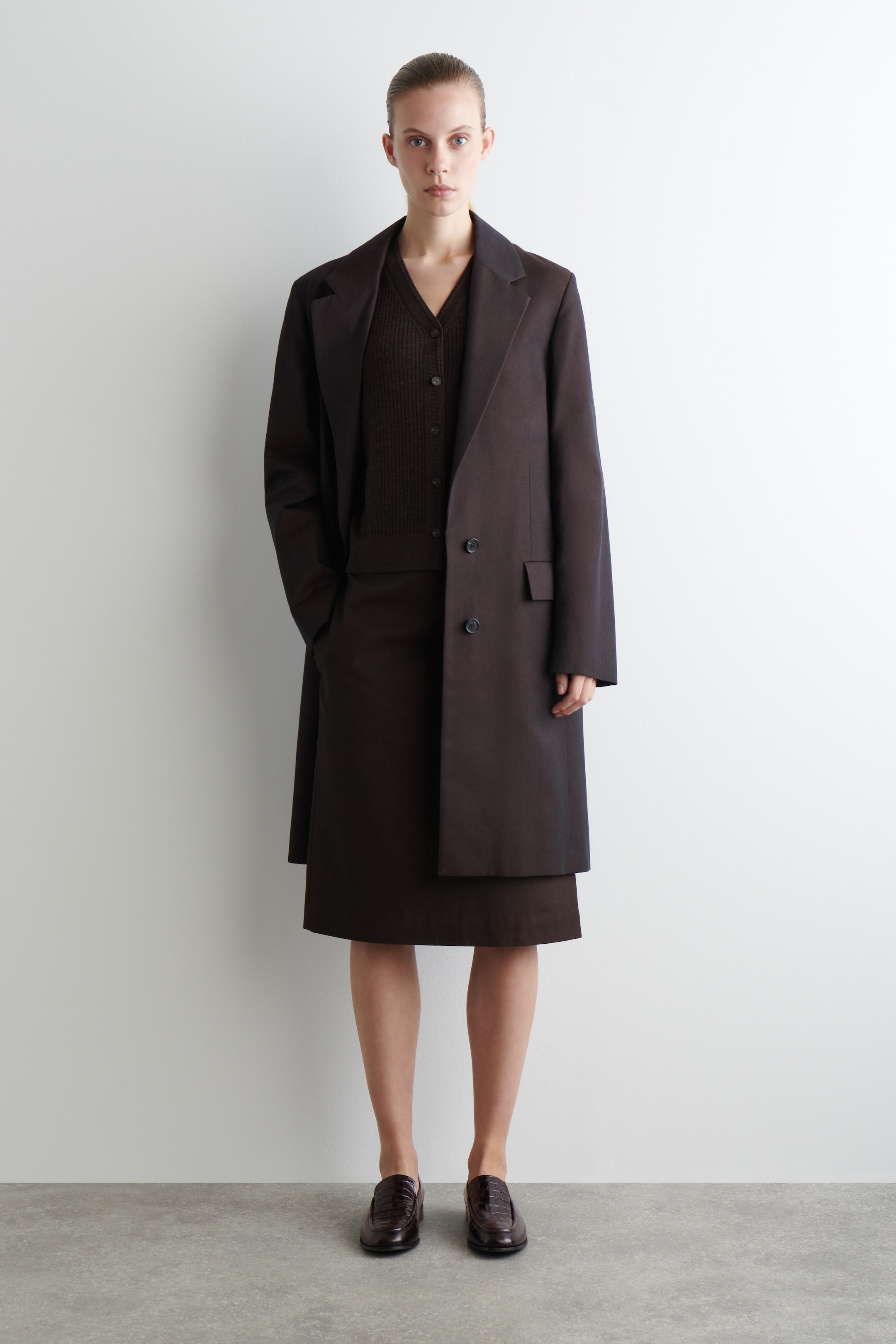 Ingrandisci l'immagine: Una vista frontale mostra un cappotto lungo marrone scuro con revers a tacca e due bottoni visibili, indossato aperto su un cardigan marrone scuro con scollo a V e bottoni sul davanti e una gonna al ginocchio. Mocassini marrone scuro con texture sono indossati.