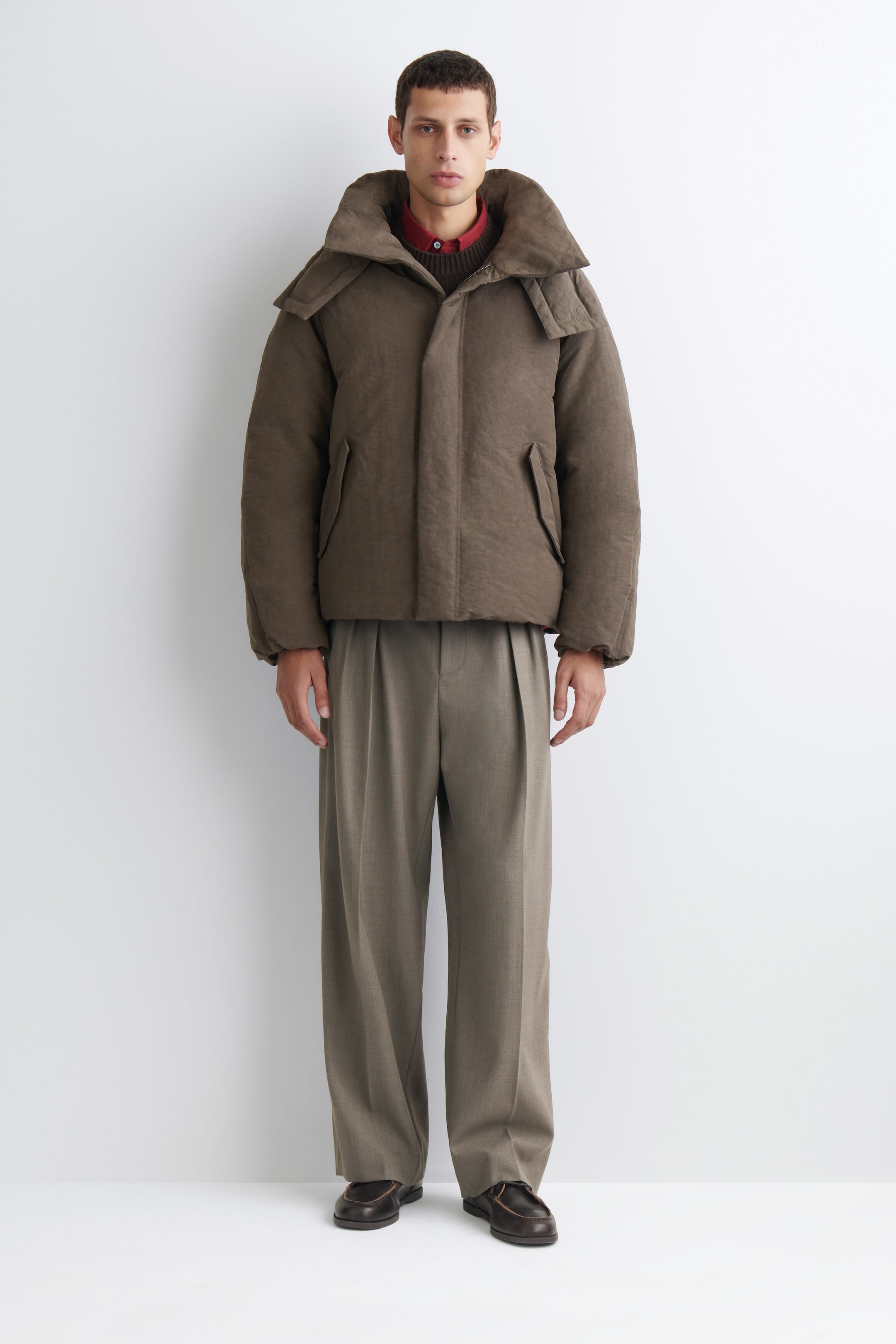 The oversized nylon-linen hooded down puffer jacket is shown from the front in a boxy, padded silhouette in a matte taupe-brown color. It features an exaggerated, high-volume hooded collar, a concealed front closure, and angled side welt pockets, layered over a dark knit sweater and a red collared shirt peeking out at the neck. The oversized nylon-linen hooded down puffer jacket is shown from the front in a boxy, padded silhouette in a matte taupe-brown color. It features an exaggerated, high-volume hooded collar, a concealed front closure, and angled side welt pockets, layered over a dark knit sweater and a red collared shirt peeking out at the neck.