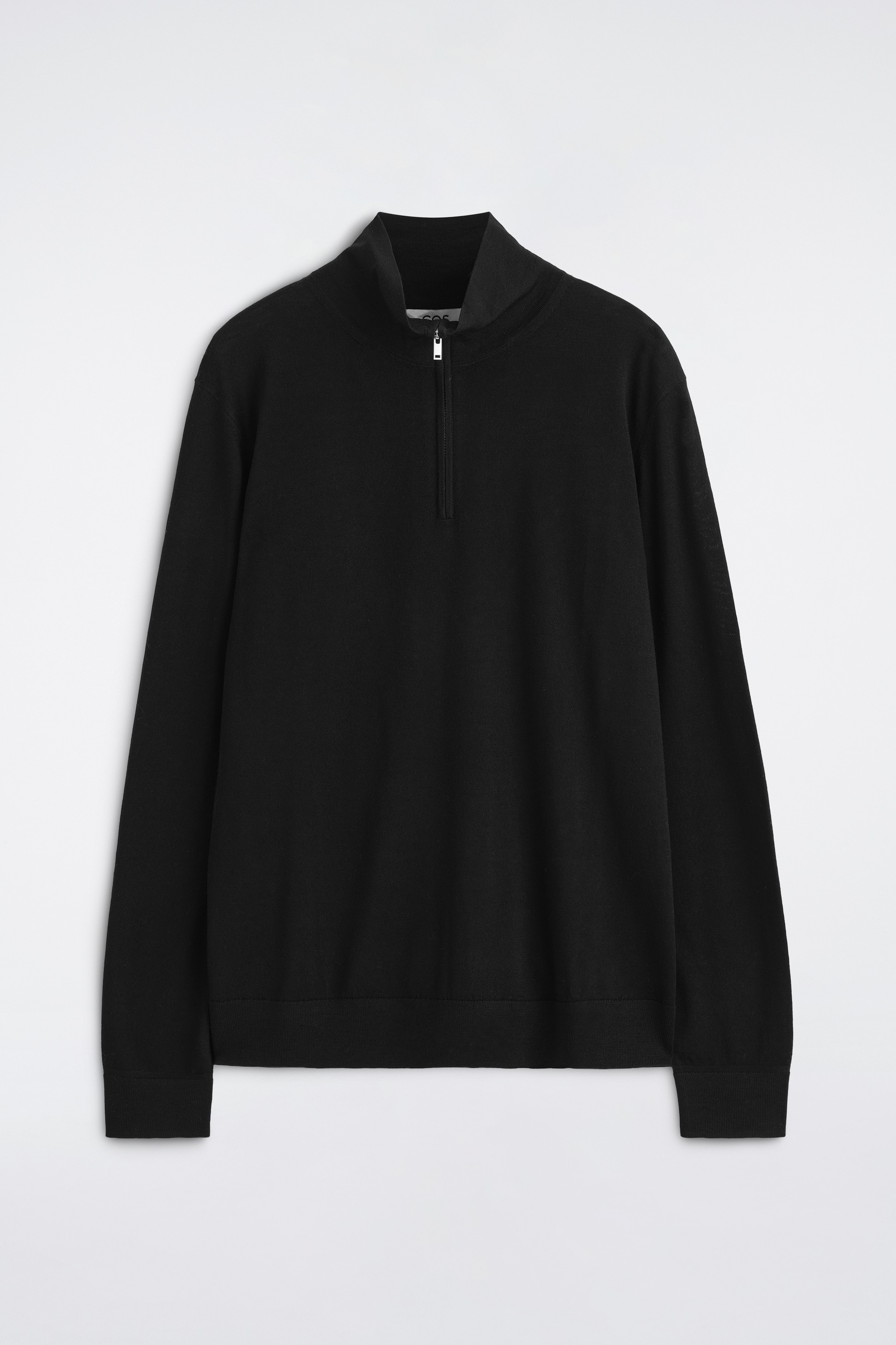 La modella indossa un maglione nero in lana merino con collo a lupetto e zip, infilato dentro pantaloni plissettati marrone scuro. Il maglione presenta una mezza zip argentata al colletto, lasciata leggermente aperta, e maniche lunghe con polsini a costine.