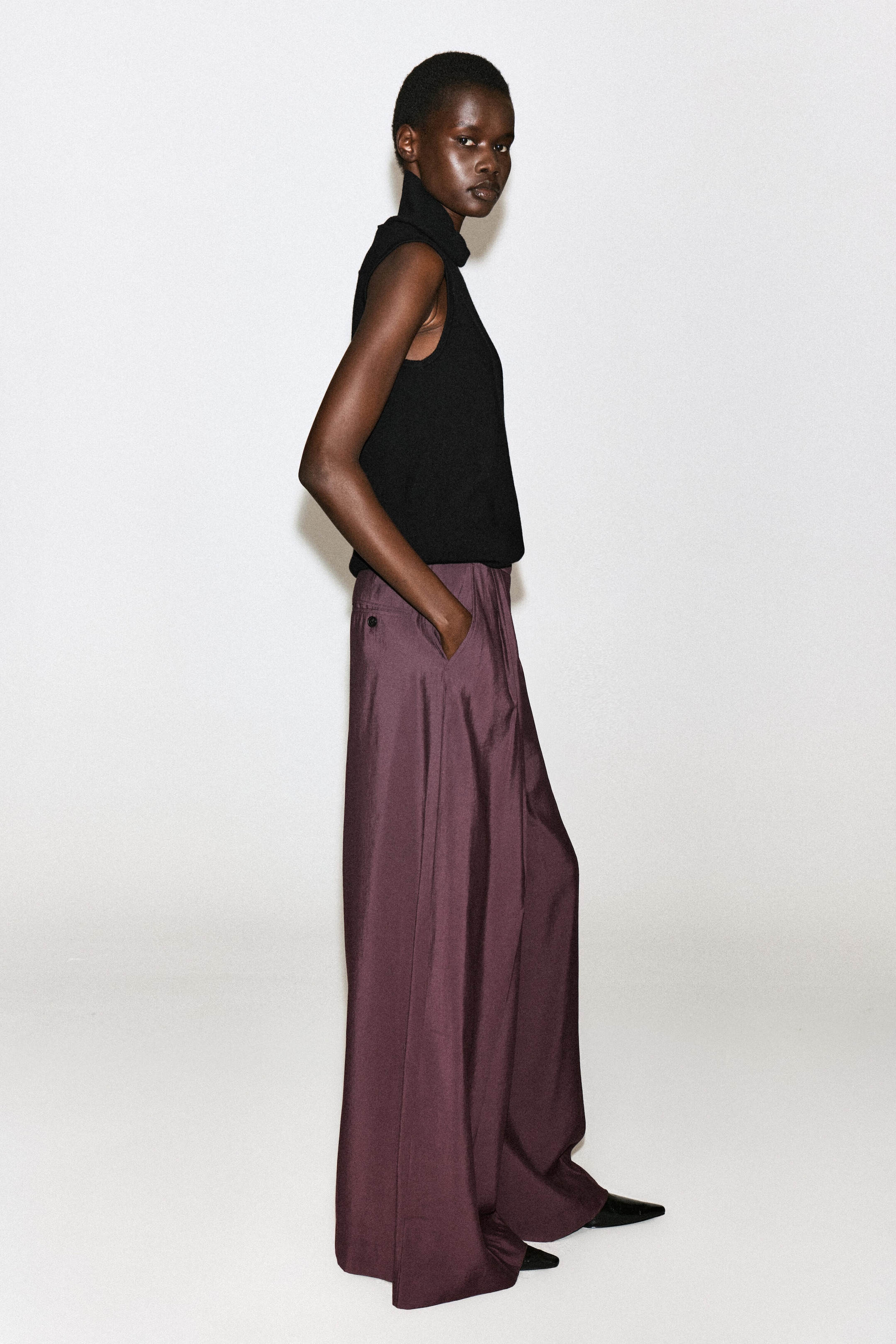Plissierte Wide-Leg-Hose aus Modal in einem tiefen Pflaumenton, getragen vom Model, das im Profil steht und eine Hand in die Seitentasche gesteckt hat. Die Hose mit lockerer Passform ist mit einem schwarzen ärmellosen Top mit hohem Ausschnitt und schwarzen Schuhen mit spitzer Zehenpartie kombiniert. Plissierte Wide-Leg-Hose aus Modal in einem tiefen Pflaumenton, getragen vom Model, das im Profil steht und eine Hand in die Seitentasche gesteckt hat. Die Hose mit lockerer Passform ist mit einem schwarzen ärmellosen Top mit hohem Ausschnitt und schwarzen Schuhen mit spitzer Zehenpartie kombiniert.