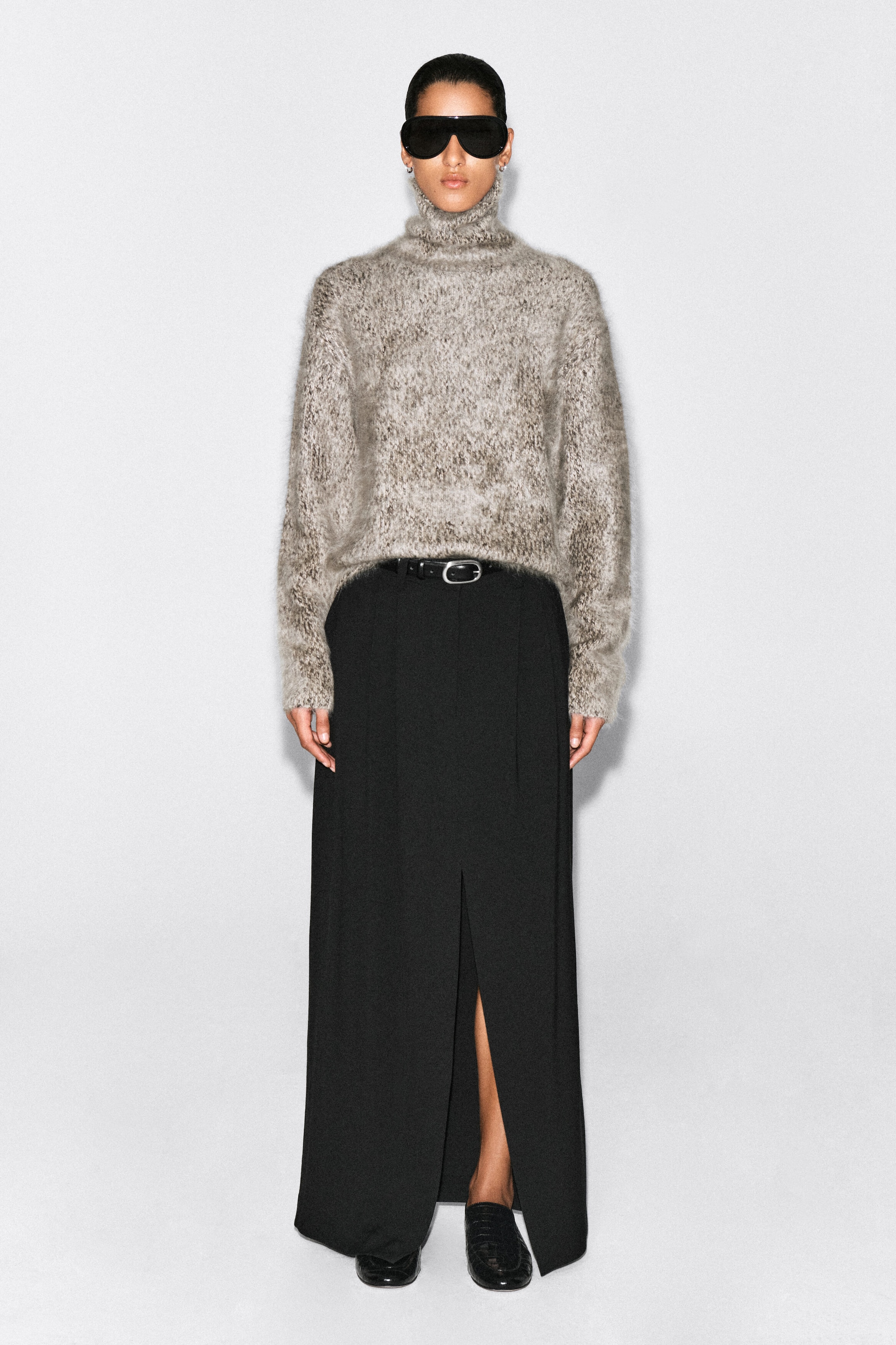 Un pull col cheminée épais, en maille chinée gris et beige, avec une texture duveteuse et brossée, est présenté de face sur un mannequin. Le pull est agencé avec une jupe longue noire fendue sur le côté et une ceinture noire. Un pull col cheminée épais, en maille chinée gris et beige, avec une texture duveteuse et brossée, est présenté de face sur un mannequin. Le pull est agencé avec une jupe longue noire fendue sur le côté et une ceinture noire.
