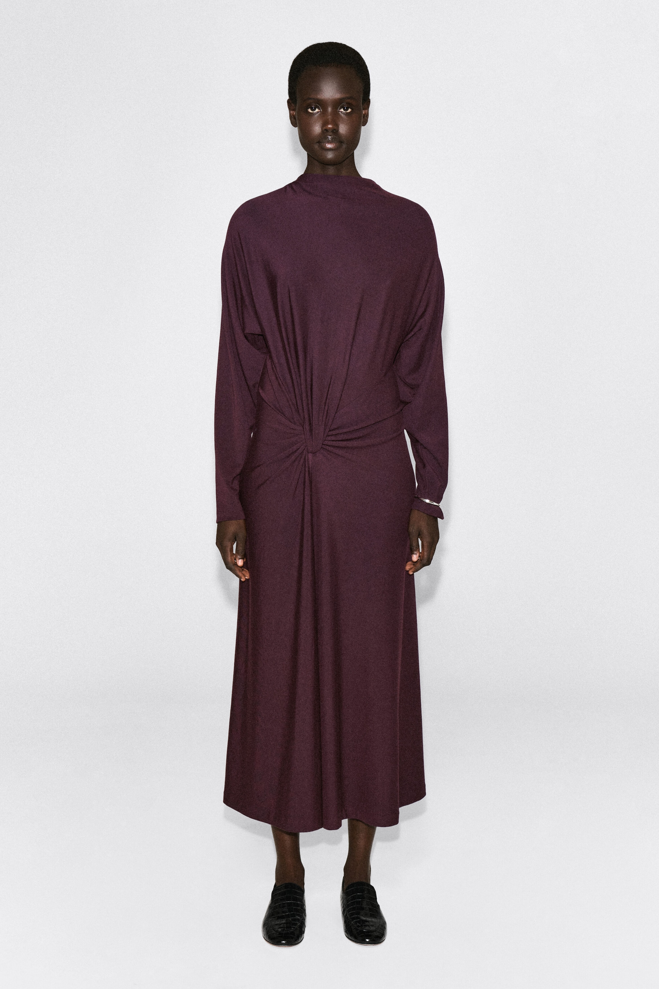 La robe midi drapée à détail torsadé dans une teinte prune profonde est présentée de face, avec un col montant et des manches longues. Un détail torsadé bien visible à la taille, au centre, crée des plis drapés et froncés qui retombent sur le corsage et le long de la jupe midi. La robe midi drapée à détail torsadé dans une teinte prune profonde est présentée de face, avec un col montant et des manches longues. Un détail torsadé bien visible à la taille, au centre, crée des plis drapés et froncés qui retombent sur le corsage et le long de la jupe midi.