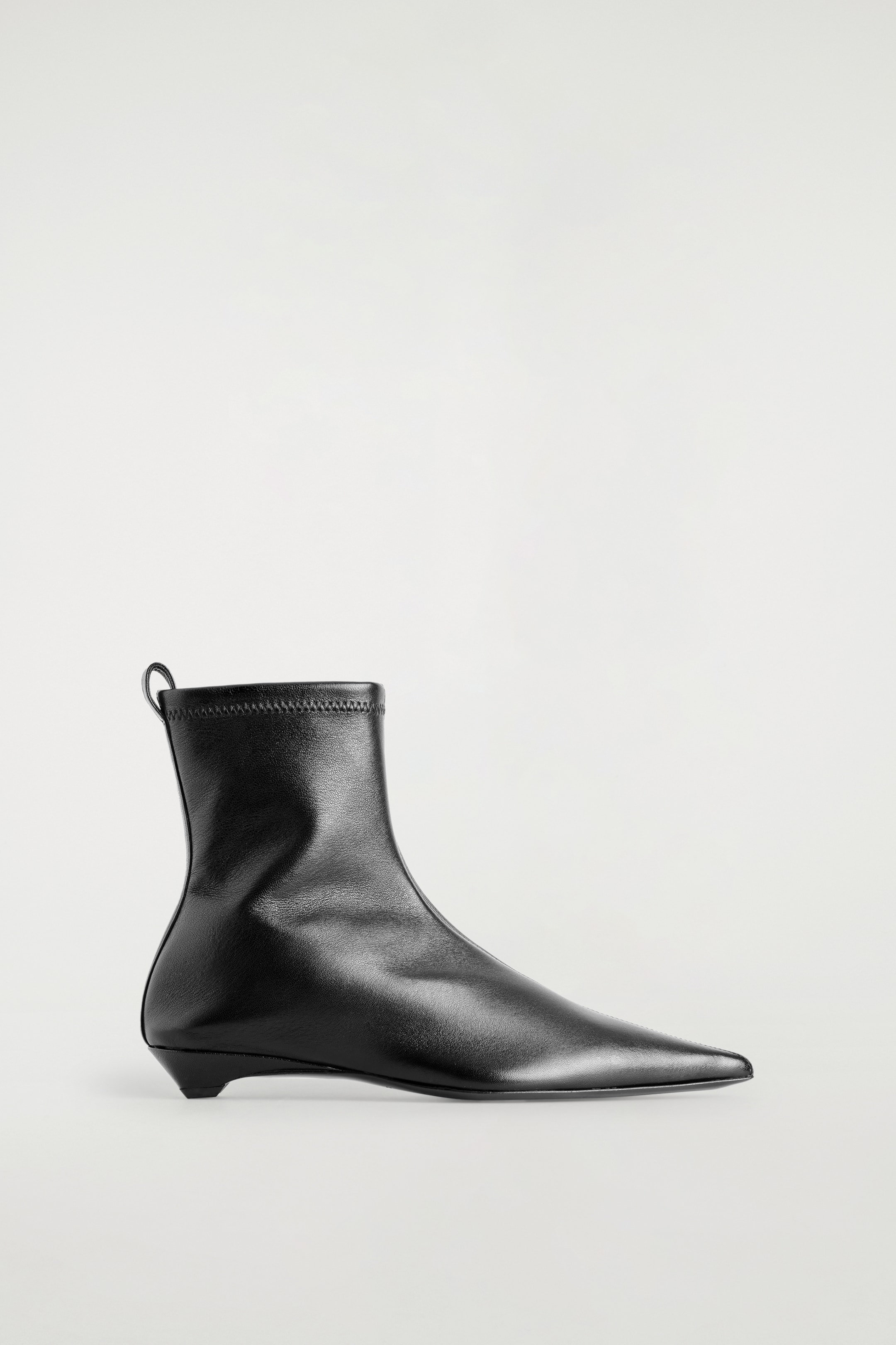 Agrandir l'image: Une botte-chaussette en cuir noir pointue est exposée, orientée vers la droite. Elle présente une tige en cuir lisse menant à un bout pointu, et une tige de cheville ajustée, semblable à une chaussette, finie avec un revers élastiqué cousu et une petite languette à l'arrière. Un talon bas et effilé soutient la botte.