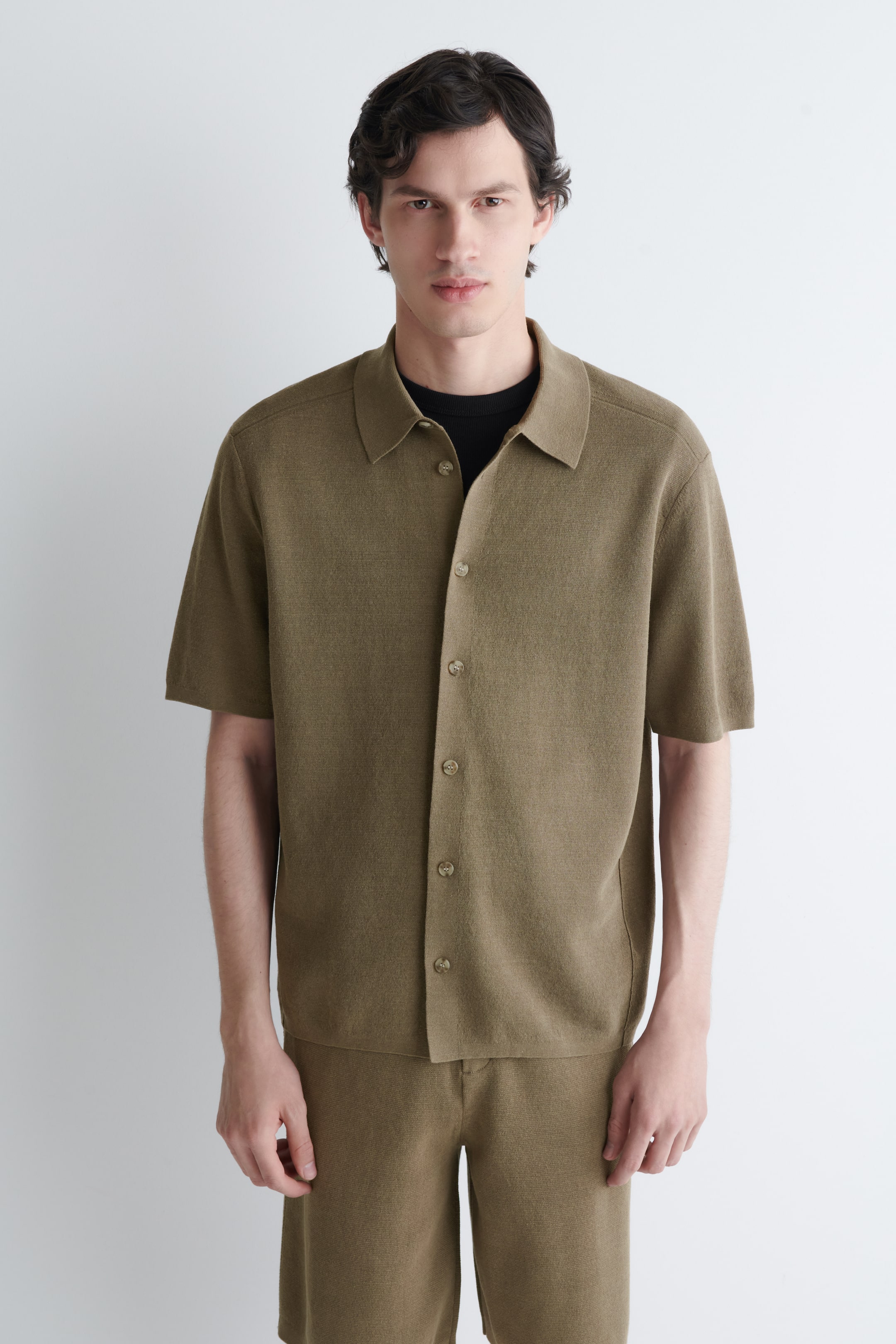 Grotere afbeelding bekijken: Een olijfgroen gebreid shirt met korte mouwen, voorzien van een klassieke kraag en een knoopsluiting aan de voorkant met lichtgekleurde knopen, wordt gedragen over een zwart t-shirt en gecombineerd met bijpassende gebreide shorts.