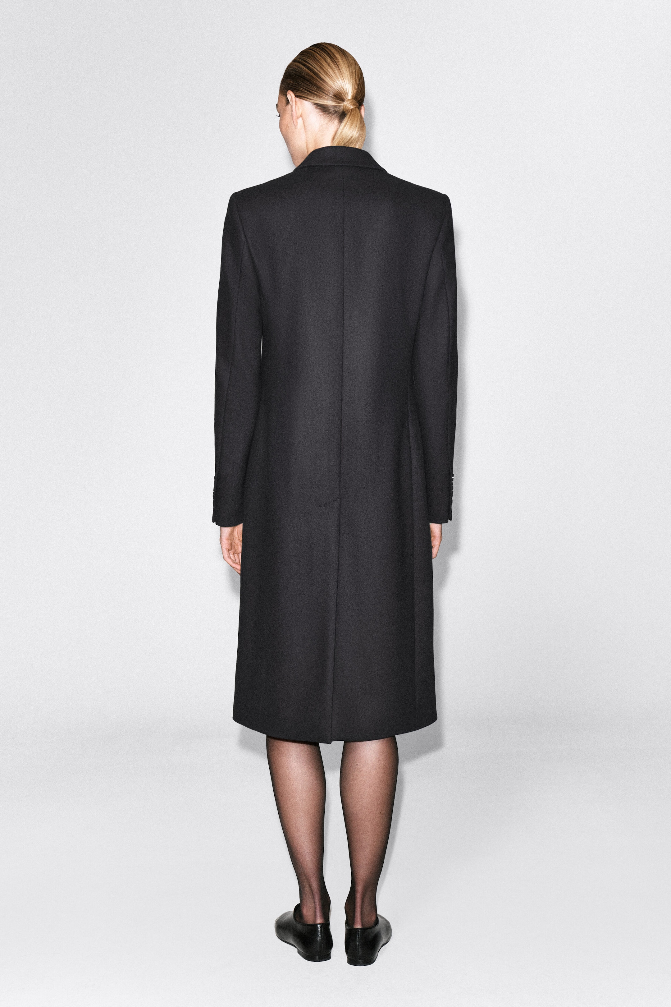 The model is wearing a long black coat and facing away to showcase the garment’s structured slim silhouette, sharp shoulders, and a center back seam with a split hem. The coat is styled with sheer black tights and flat black shoes. The model is wearing a long black coat and facing away to showcase the garment’s structured slim silhouette, sharp shoulders, and a center back seam with a split hem. The coat is styled with sheer black tights and flat black shoes.