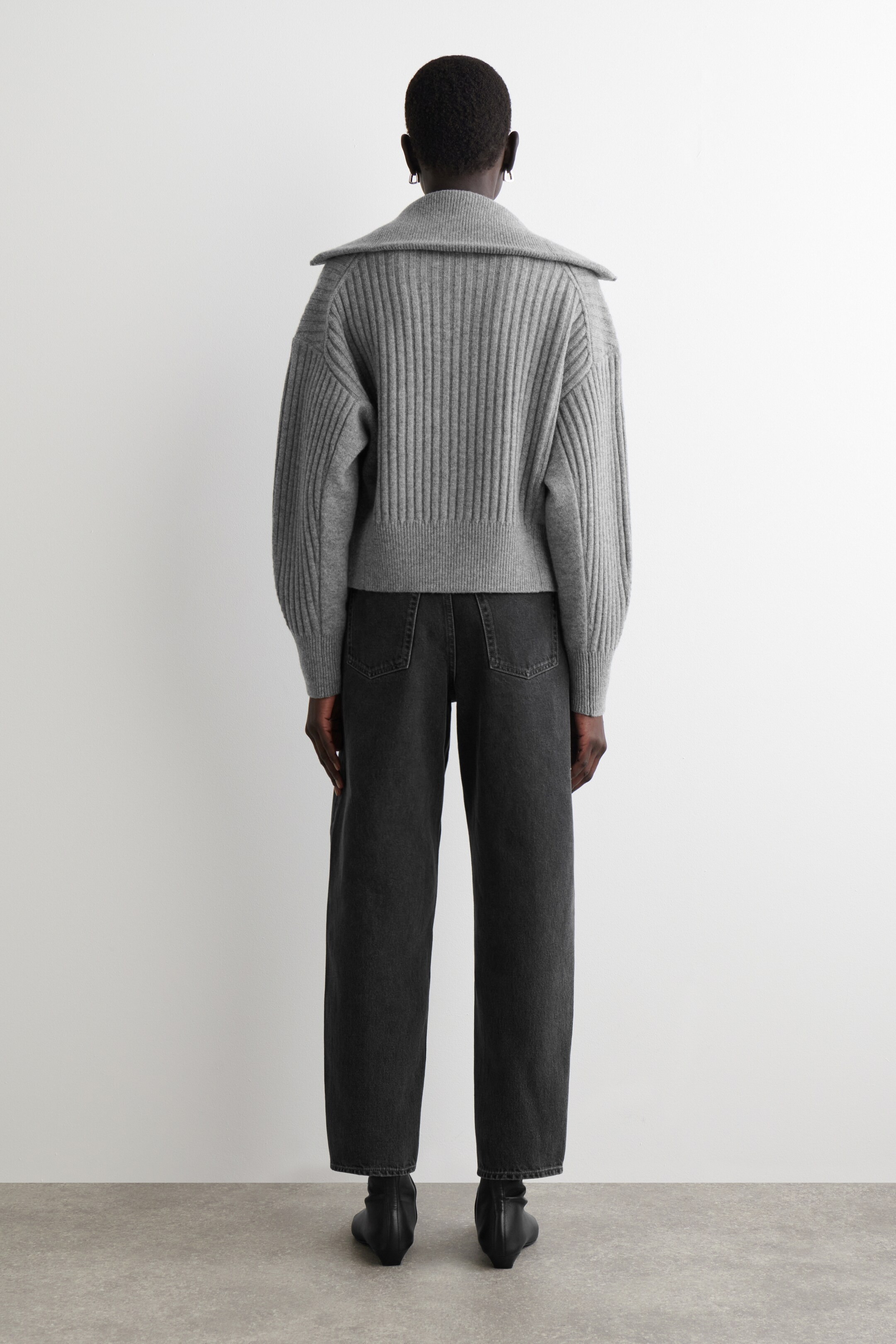Größeres Bild anzeigen: Ein grauer gerippter Strickpullover wird von hinten gezeigt, mit einem breiten, umgeschlagenen Kragen, einem vertikal gerippten Körper und Ärmeln, gerippten Bündchen und einem kurzen gerippten Saum.