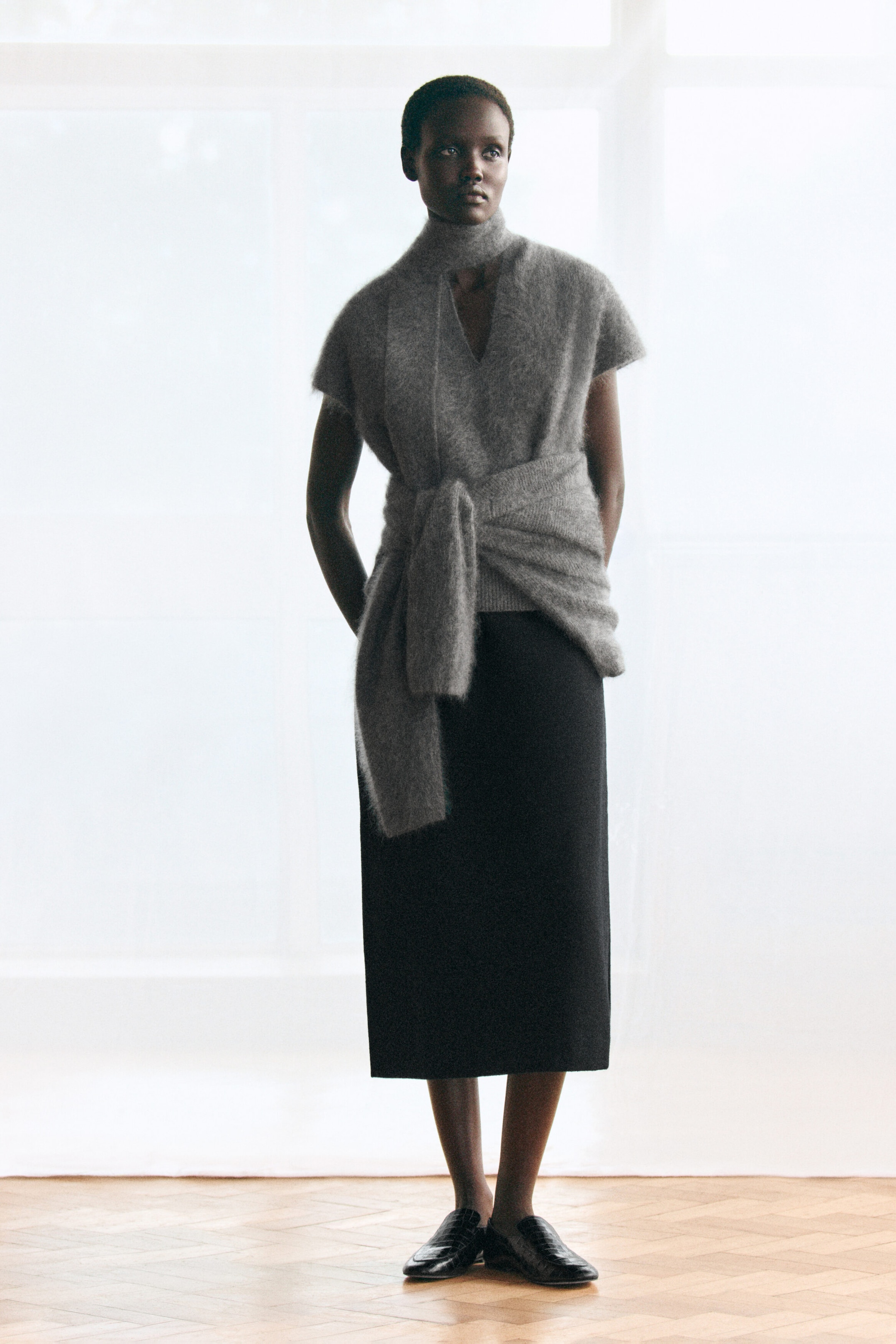 Le haut en laine mérinos brossée gris drapé est présenté de face, avec une maille texturée et duveteuse, un col en V et de courtes manches cap. Une écharpe en maille assortie s’enroule autour du cou et se noue à la taille, créant un effet superposé et drapé par-dessus une jupe midi noire. Le haut en laine mérinos brossée gris drapé est présenté de face, avec une maille texturée et duveteuse, un col en V et de courtes manches cap. Une écharpe en maille assortie s’enroule autour du cou et se noue à la taille, créant un effet superposé et drapé par-dessus une jupe midi noire.