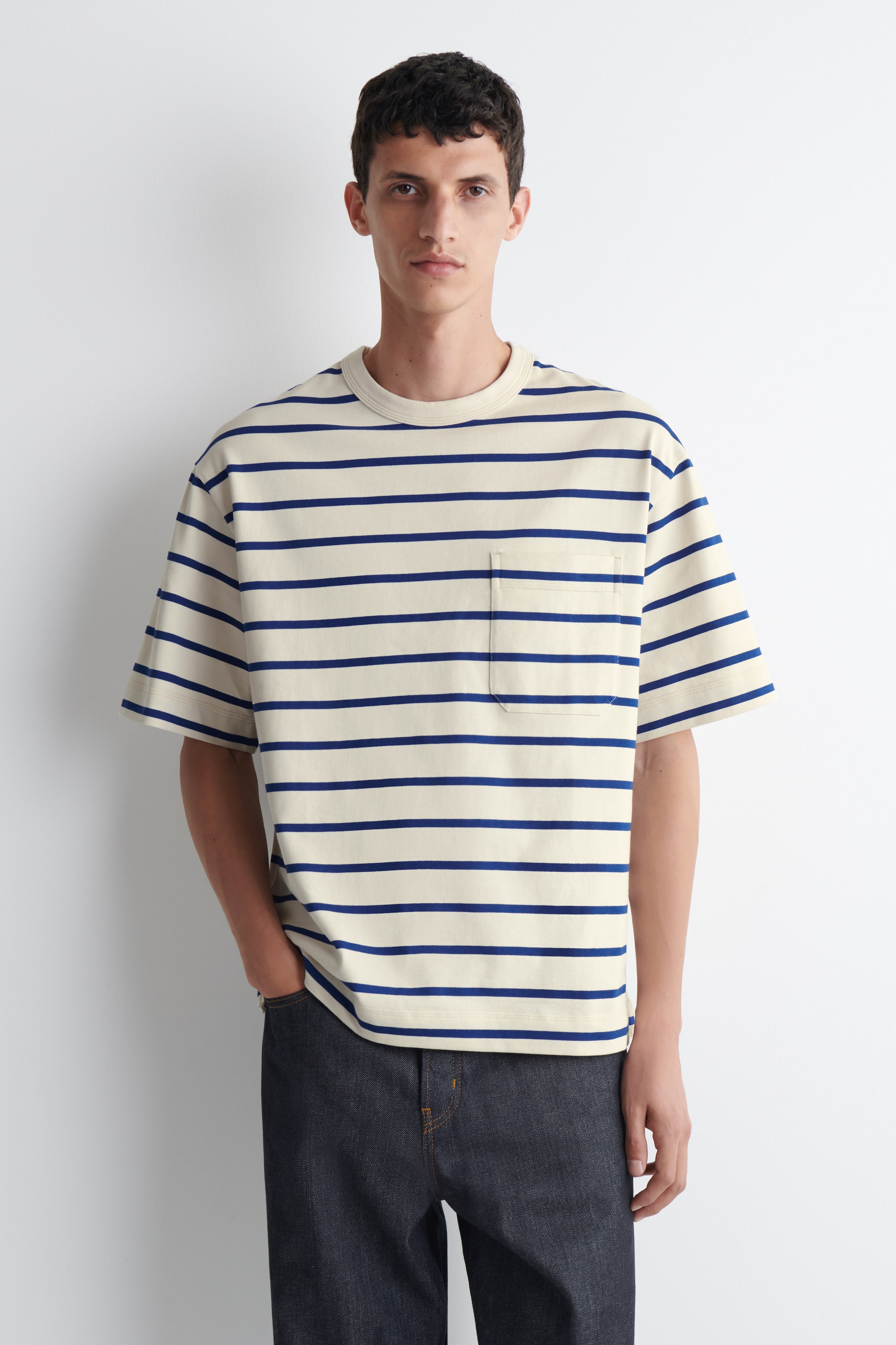 Grotere afbeelding bekijken: Een crèmekleurig T-shirt met horizontale marineblauwe strepen wordt van voren getoond, met een geribde ronde hals, korte mouwen en een borstzakje links op de borst.